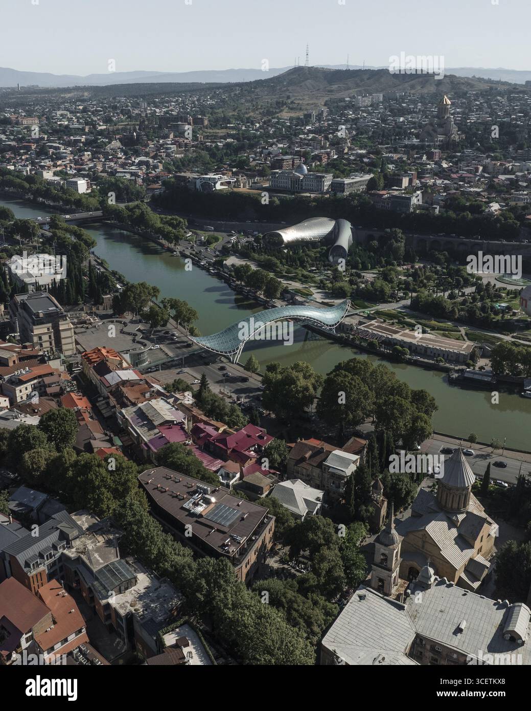 Vista aerea del fiume tortuoso che riflette il cielo, degli edifici storici con tetti rossi e del moderno Peace Bridge che contrasta con l'antica fortezza di Narikala, Tbilisi, Tbilisi, Georgia. Foto Stock