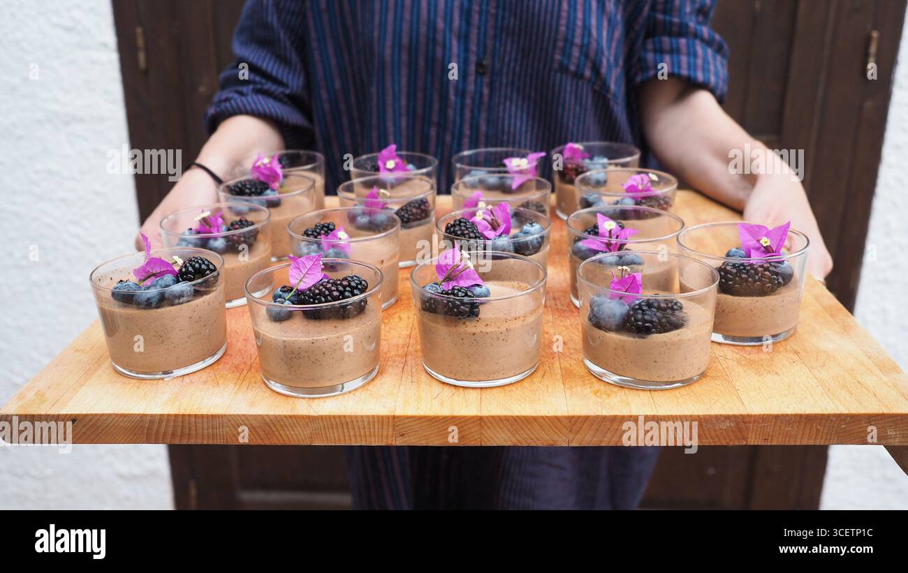 Persona che tiene un vassoio di legno con budini di cioccolato a base di more, mirtilli e fiori commestibili. Foto Stock