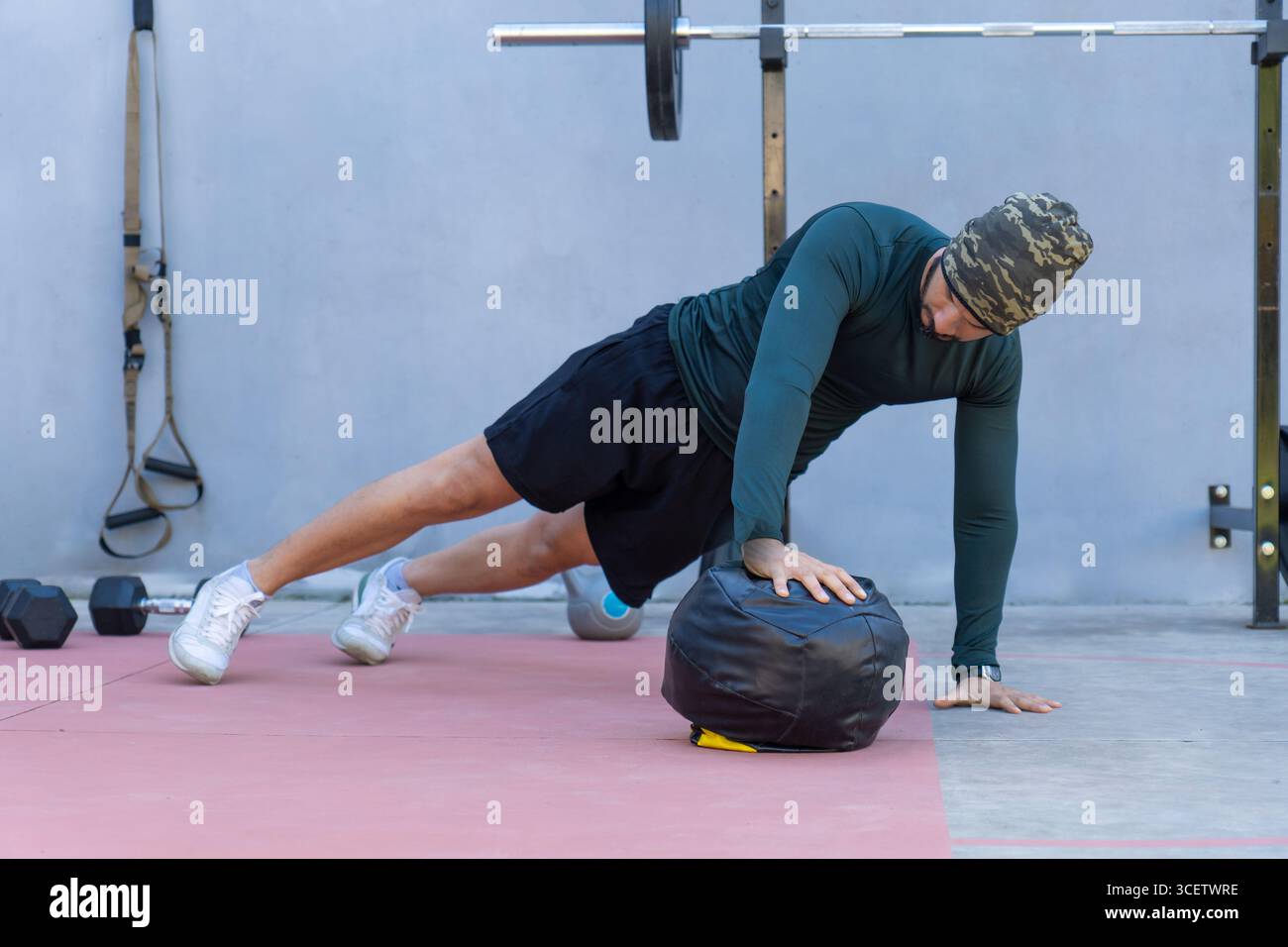 Atleta maschile che indossa un berretto che esegue push-up con una sola mano su una palla medica in una palestra all'aperto, dimostrando forza e dedizione al fitness Foto Stock