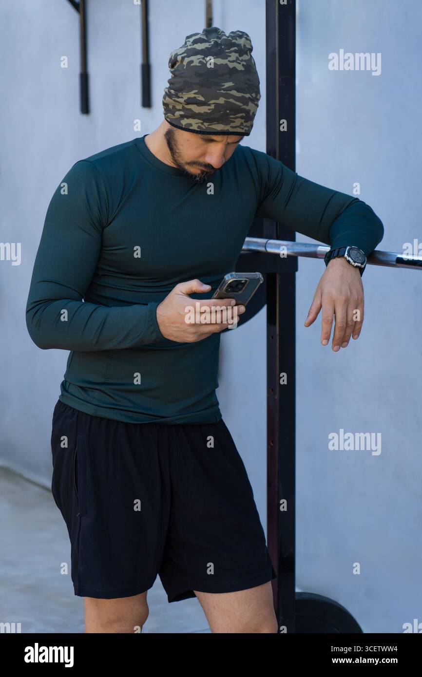 L'atleta che indossa un berretto mimetico, una camicia a maniche lunghe e pantaloncini corti controlla il suo smartphone dopo una sessione di allenamento in una palestra all'aperto, Foto Stock