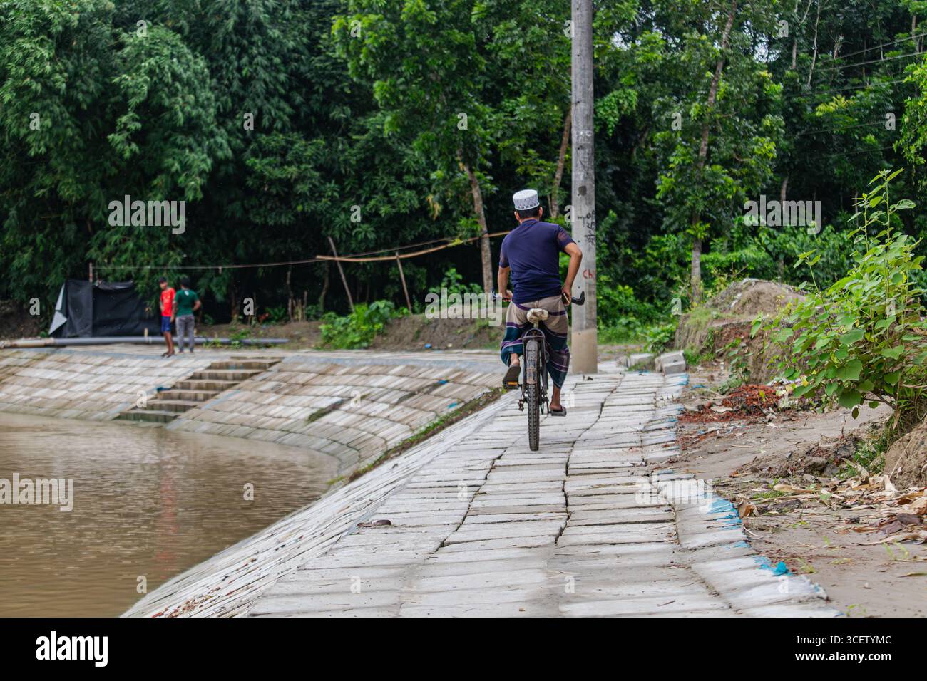 Uomo del posto che pedala lungo l'argine lastricato accanto all'acqua di alluvione marrone sotto alberi verdi del villaggio Foto Stock