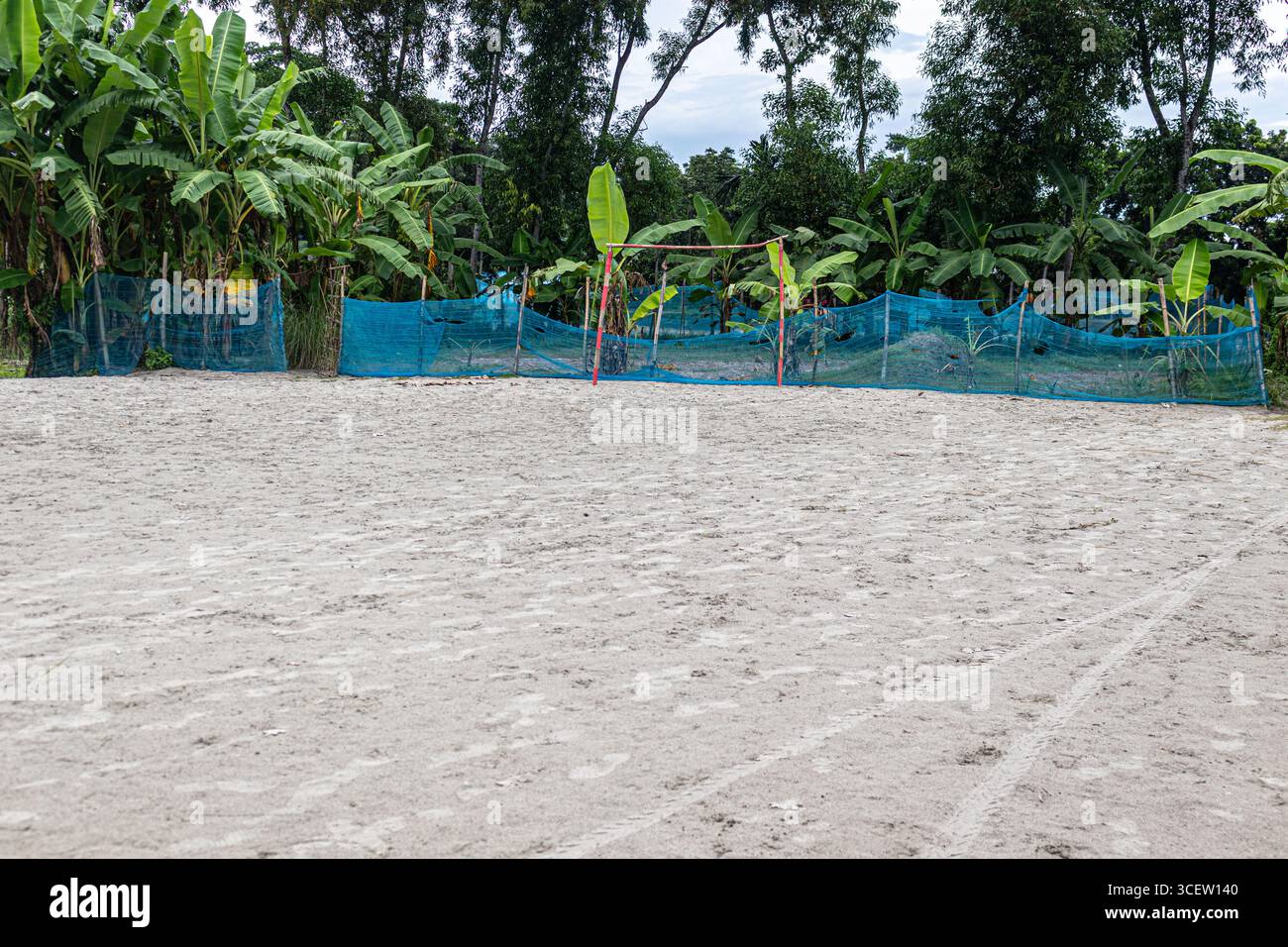 Ampio campo aperto sabbioso per partite di calcio per bambini delimitato da piante tropicali di banane e recinzione a rete blu Foto Stock