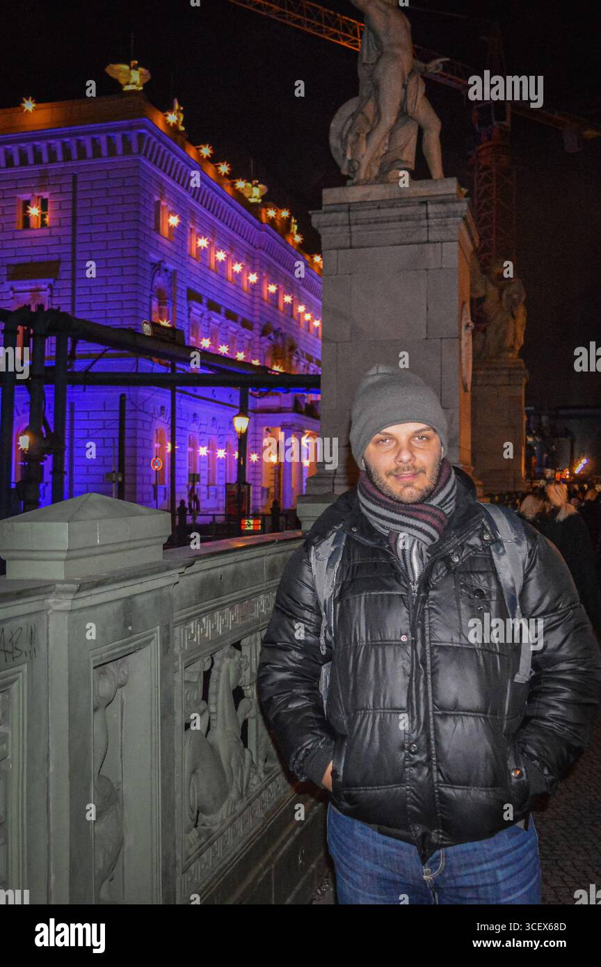 Uomo in giacca e sciarpa invernale di notte sul Ponte Schloss di Berlino con edificio illuminato e statue sullo sfondo. Foto Stock