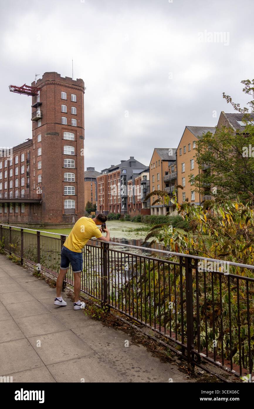 Un uomo appoggiato su una ringhiera e che scatta una foto lungo un passaggio pedonale lungo il fiume, con edifici storici in mattoni e appartamenti moderni sullo sfondo. Urbano Foto Stock