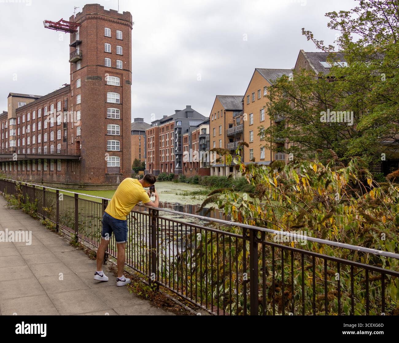 Un uomo appoggiato su una ringhiera e che scatta una foto lungo un passaggio pedonale lungo il fiume, con edifici storici in mattoni e appartamenti moderni sullo sfondo. Urbano Foto Stock