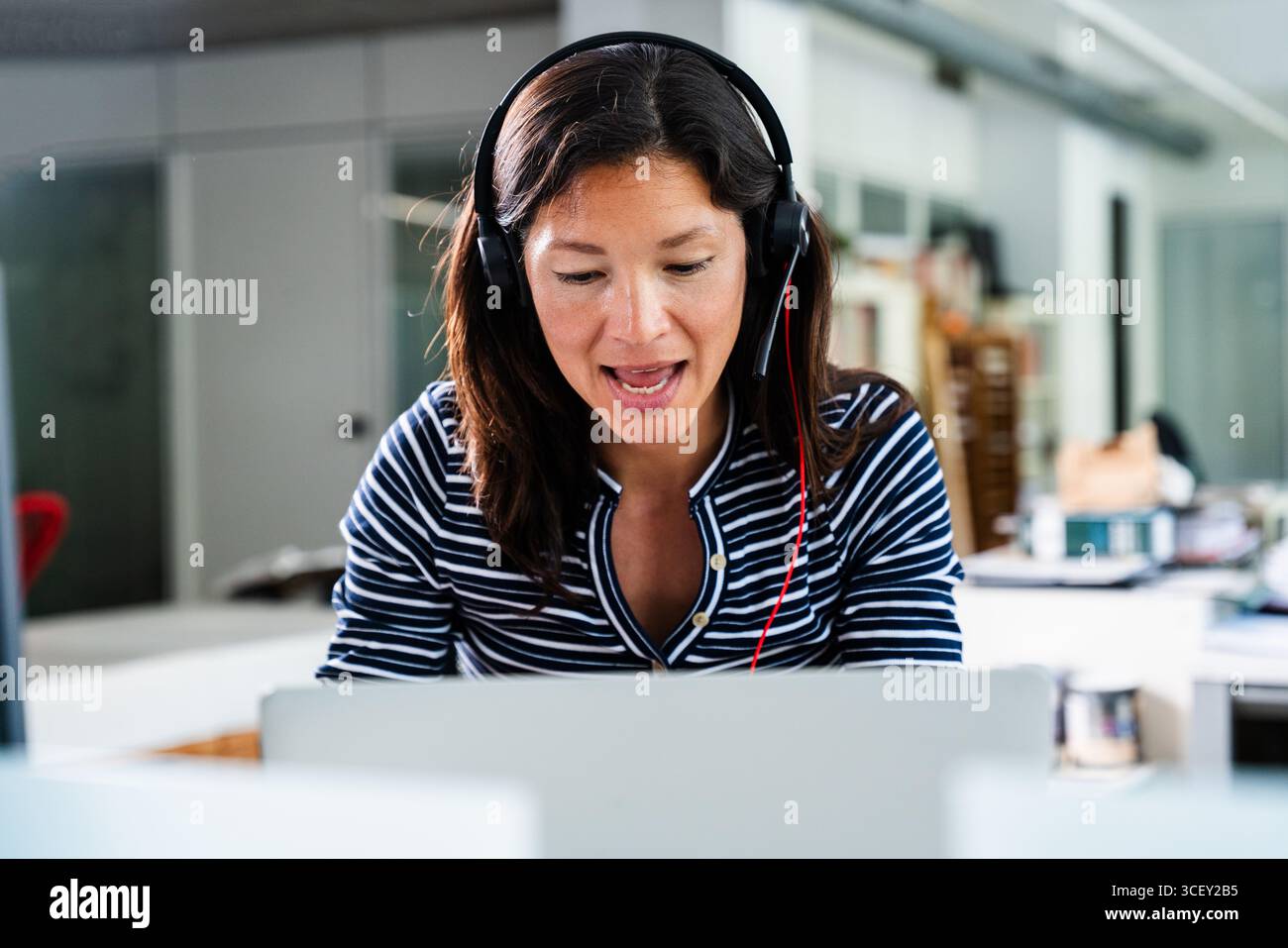 Donna d'affari concentrata che indossa una cuffia/auricolare partecipa a una videochiamata mentre lavora in remoto dal suo ufficio domestico, utilizzando un laptop per una c Foto Stock