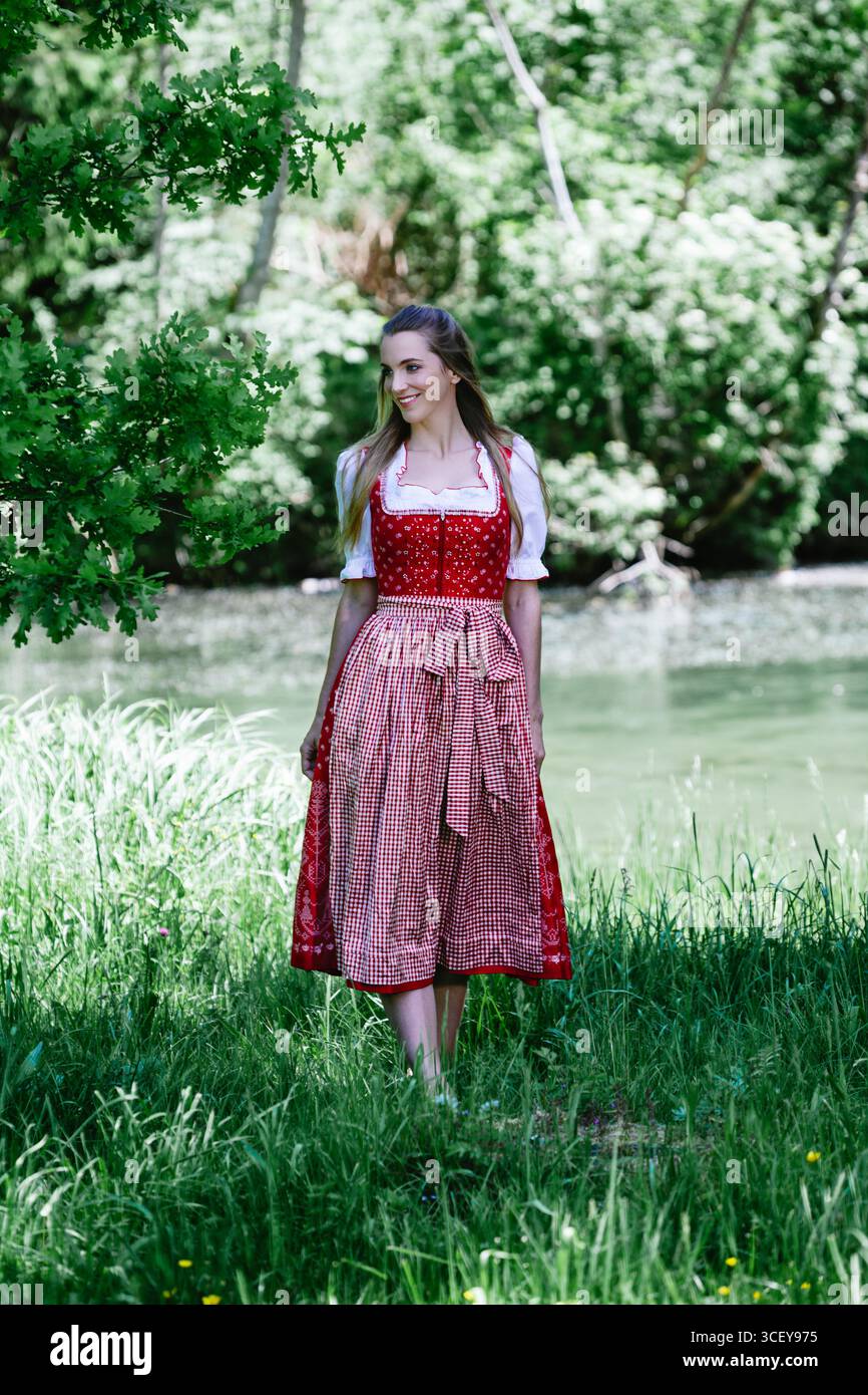 Un ritratto a figura intera di una bella giovane donna con lunghi capelli marroni, che indossa un tradizionale dirndl rosso con una camicetta bianca e un assegno rosso e bianco Foto Stock