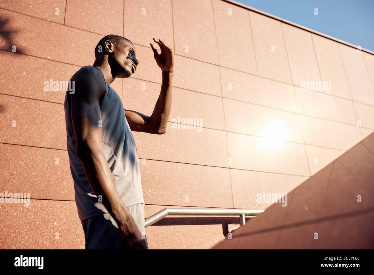 Una persona che indossa una camicia senza maniche si trova all'aperto sotto la luce del sole, proteggendo la sua visione mentre si affaccia su una facciata moderna dell'edificio. Rif. Scena Foto Stock