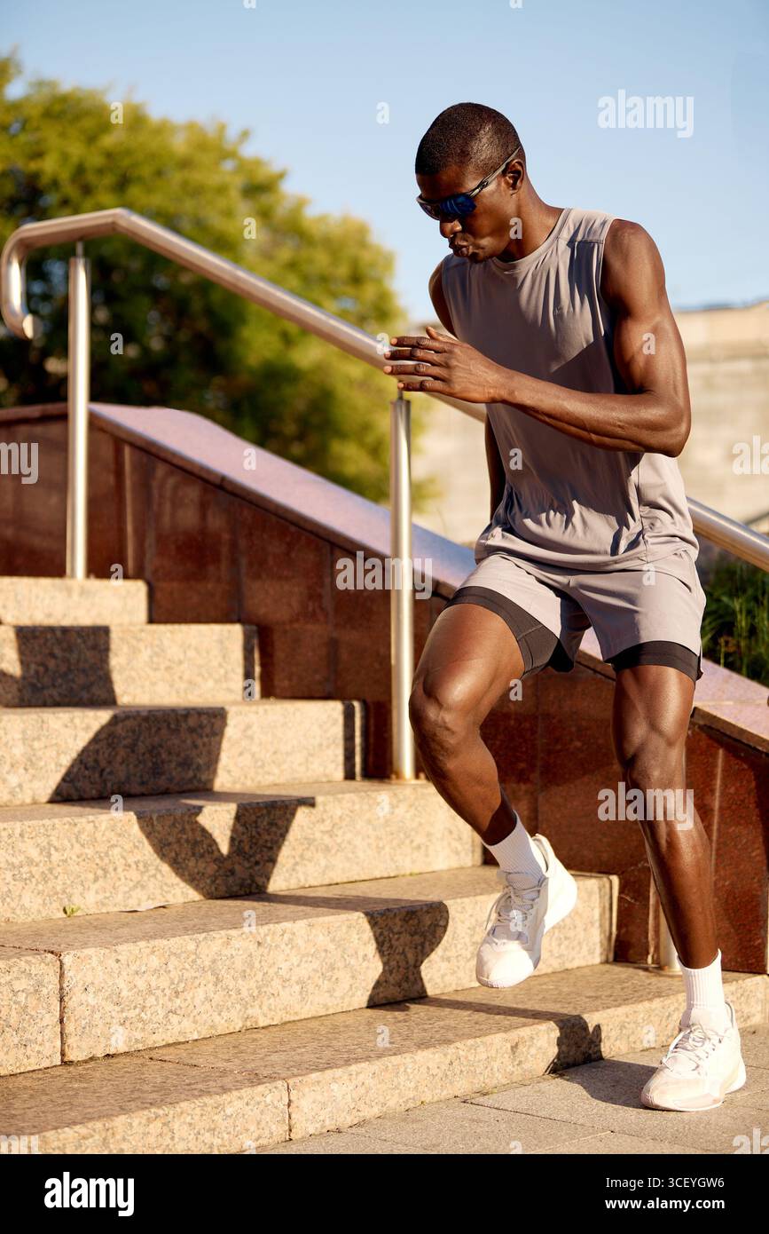 Giovane che esegue esercizi di scale per il fitness in un ambiente soleggiato all'aperto. Indossa una camicia senza maniche, pantaloncini e scarpe sportive. L'attività tak Foto Stock