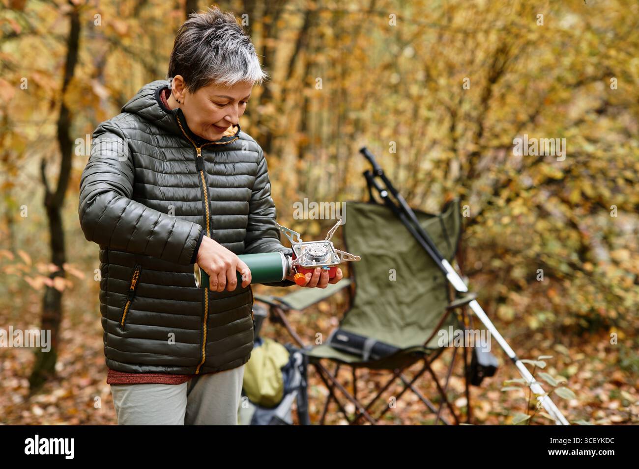 Una donna anziana prepara un pasto caldo nel suo campeggio durante una tranquilla escursione nella foresta. Foto Stock
