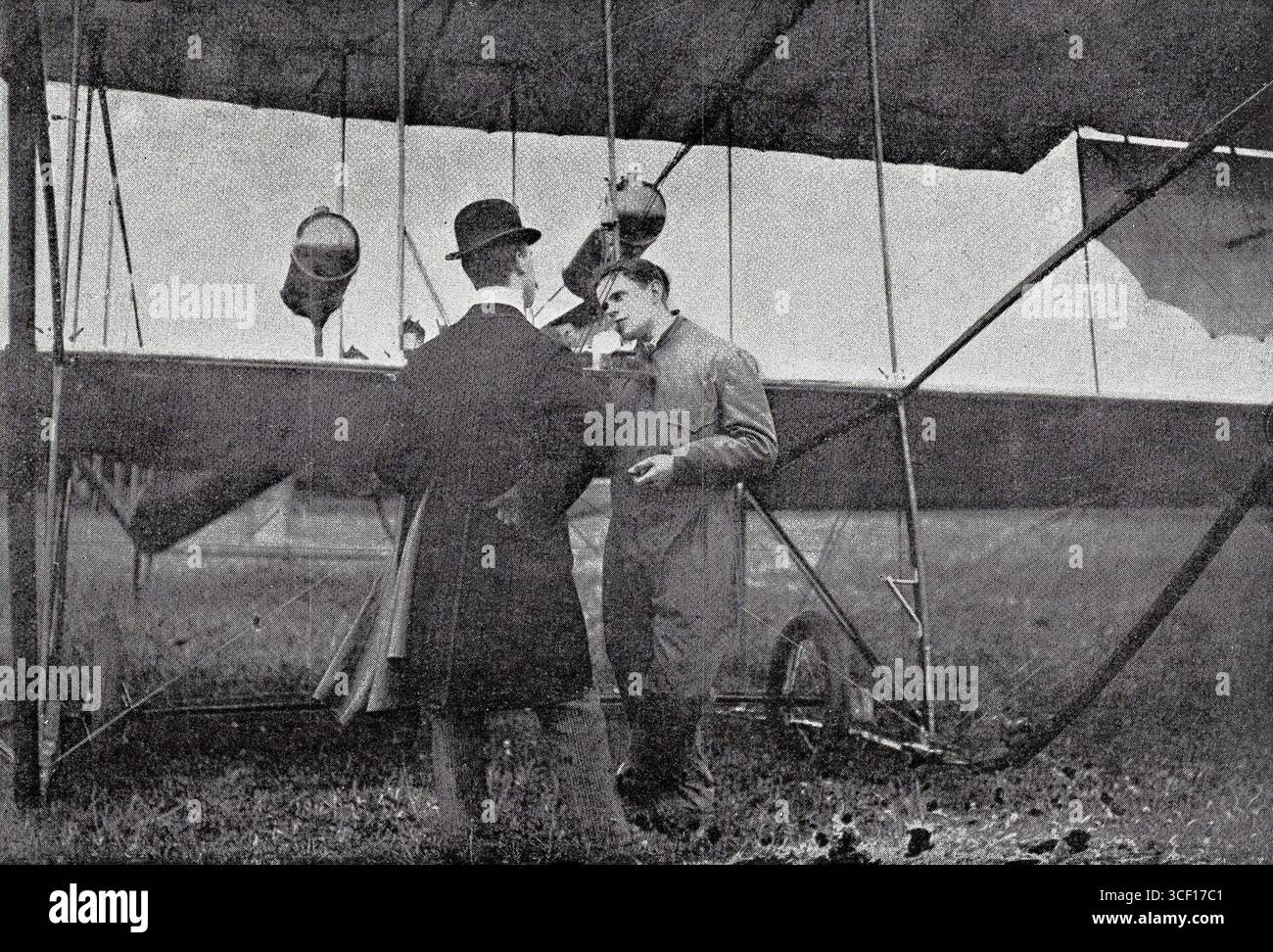 Un pilota, probabilmente intorno al 1910 nei Paesi Bassi, interagisce con un uomo in abbigliamento formale vicino al suo primitivo biplano dopo un volo. Questo momento cattura l'aviazione precoce e l'interazione sociale dei pionieri dell'aviazione. Foto Stock