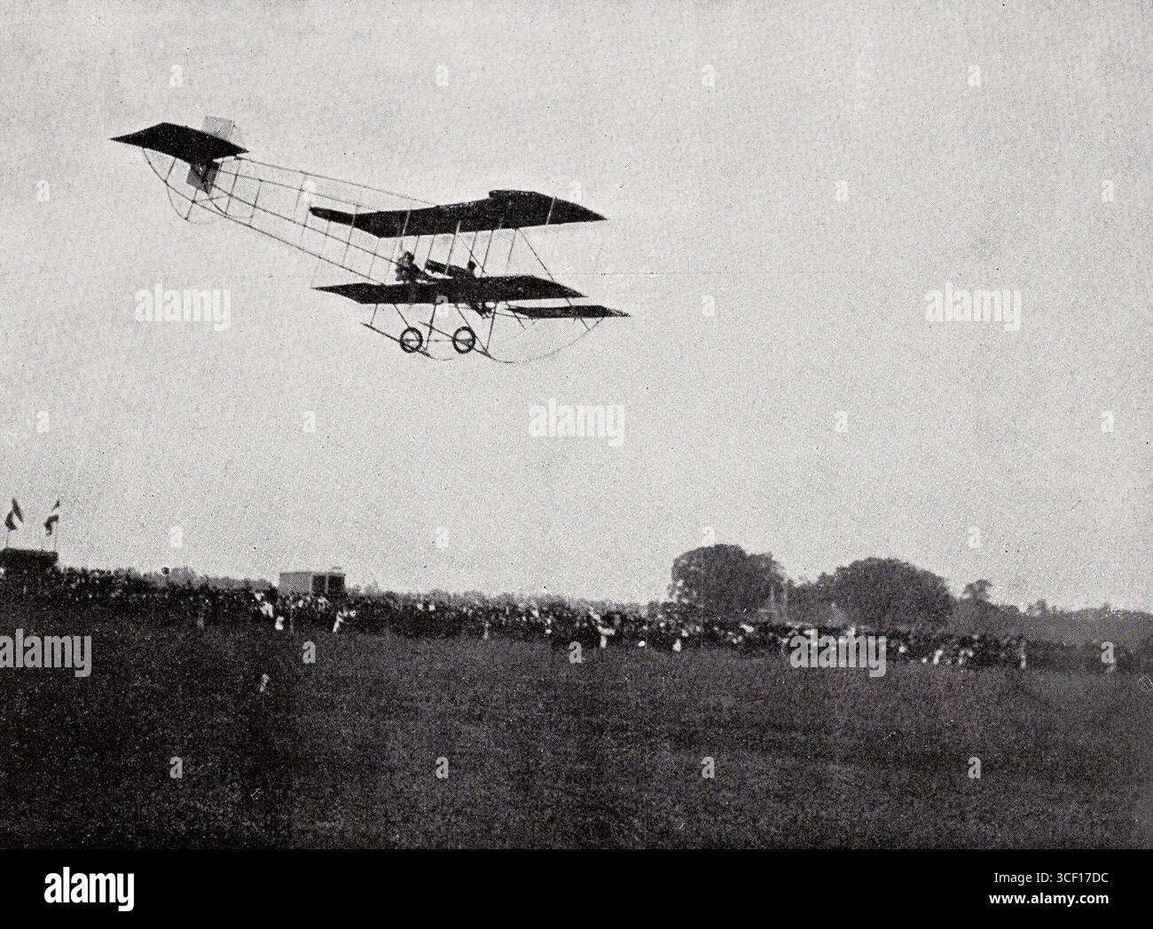 Un biplano primitivo dei primi anni '1900 è visto volare sopra un campo con spettatori. L'immagine, probabilmente di circa 1910 nei Paesi Bassi, mostra l'aviazione precoce e l'interesse pubblico per il volo. Foto Stock