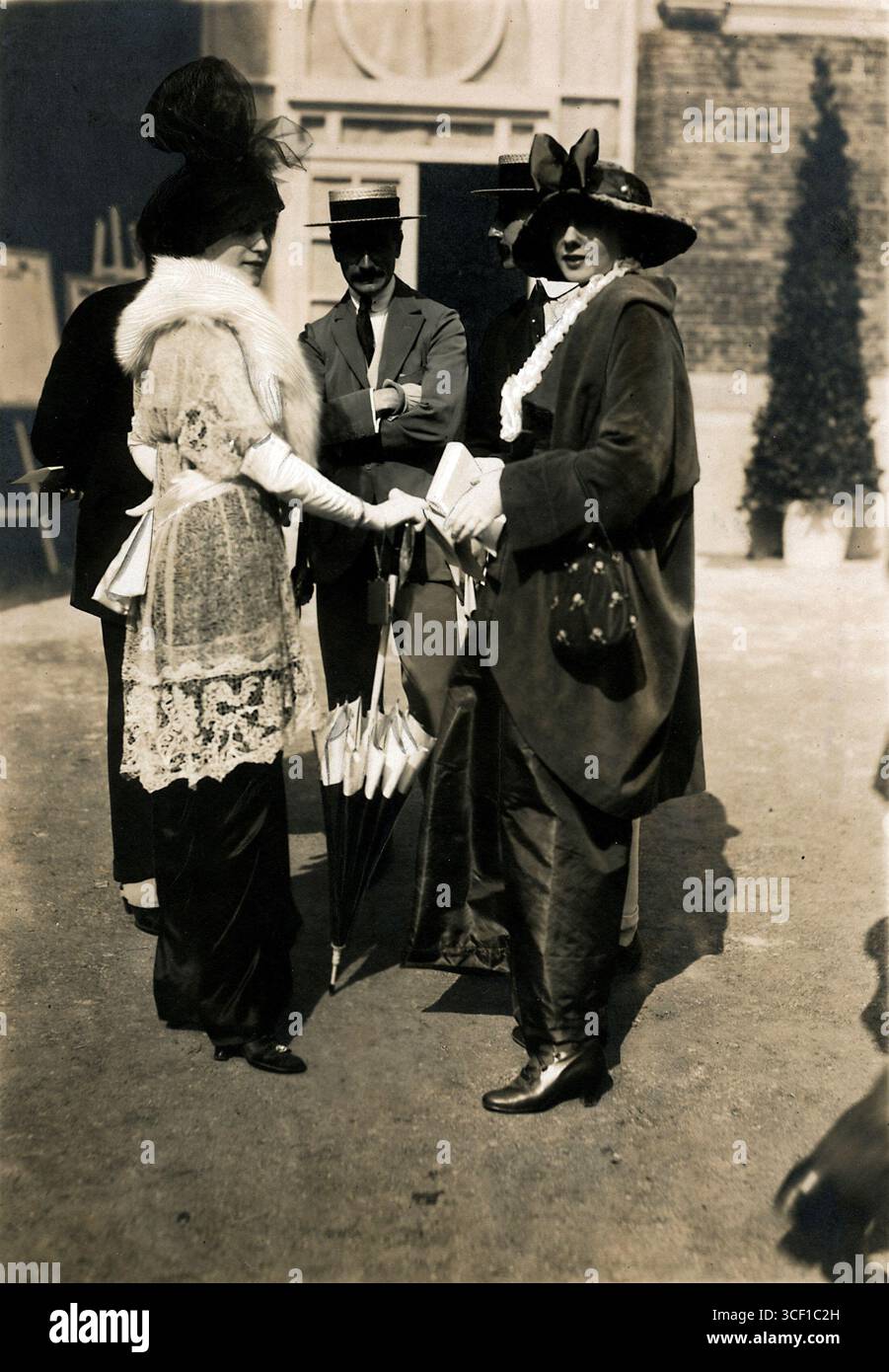 Una fotografia di Deauville, Francia, nel 1913, che mostra nuovi outfit da donna per la stagione, tra cui una tunica in pizzo, un cappello, una sciarpa di pelliccia, guanti e ombrellone. Gli uomini sullo sfondo indossano abiti con calze e cappelli di paglia che riflettono la moda del tempo. Foto Stock