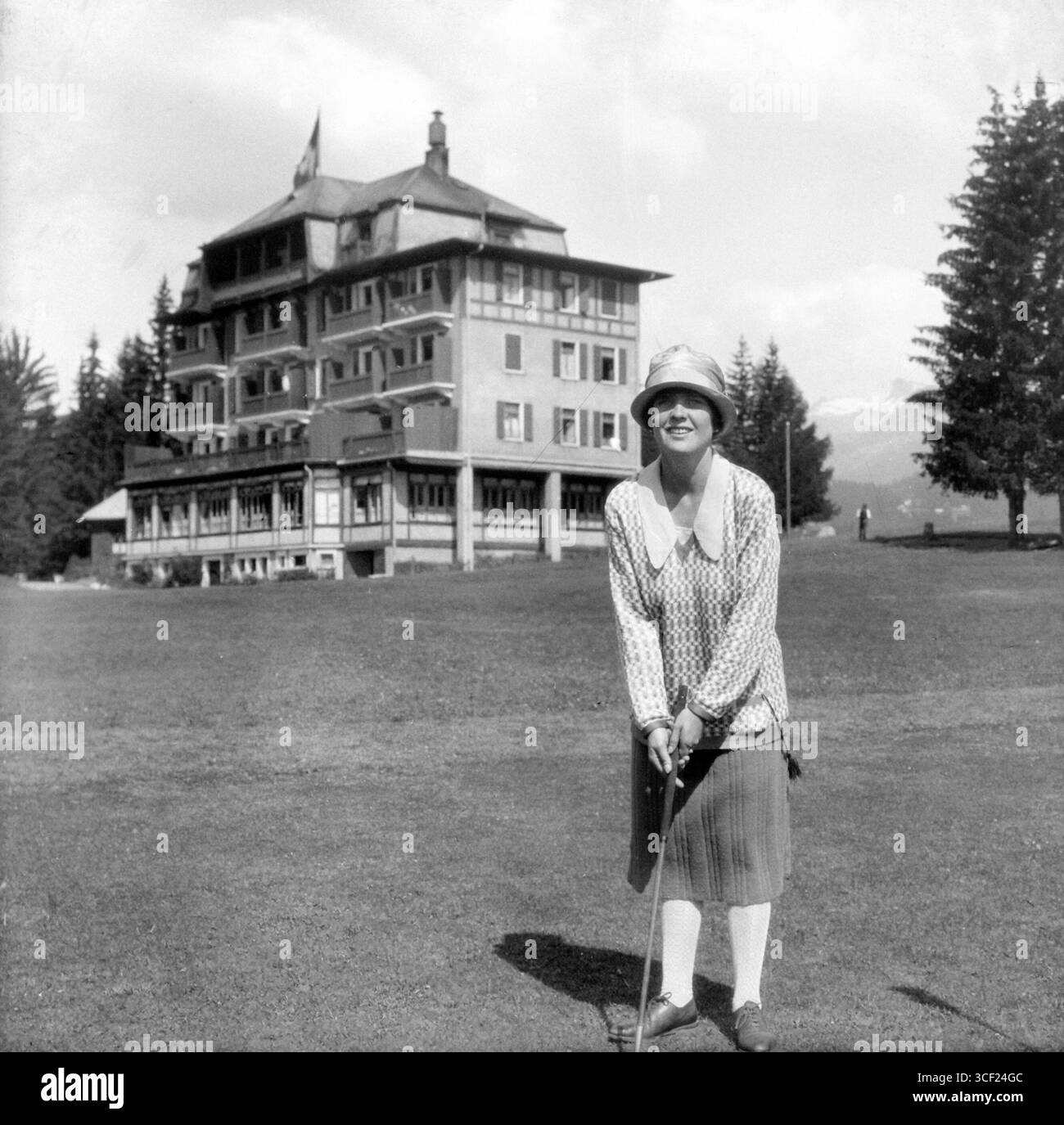 Una donna, vestita con un cappello, gioca a golf intorno al 1927. La foto, probabilmente scattata in Svizzera, mostra un hotel con una bandiera svizzera sullo sfondo. Foto Stock