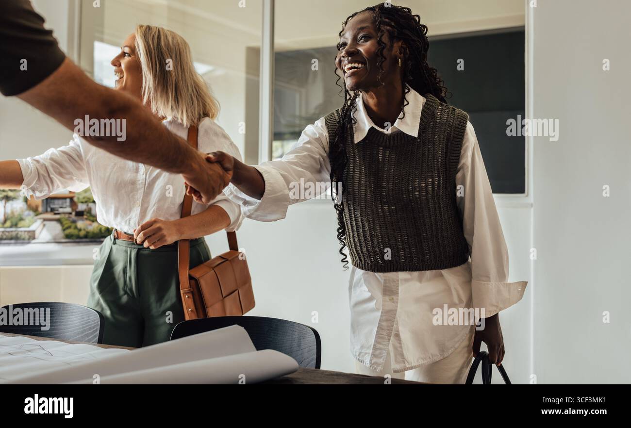 Due architetti donna, una giovane adulta e una di mezza età, stringono la mano a un cliente nel loro ufficio moderno, simboleggiando il lavoro di squadra e la collaborazione professionale Foto Stock