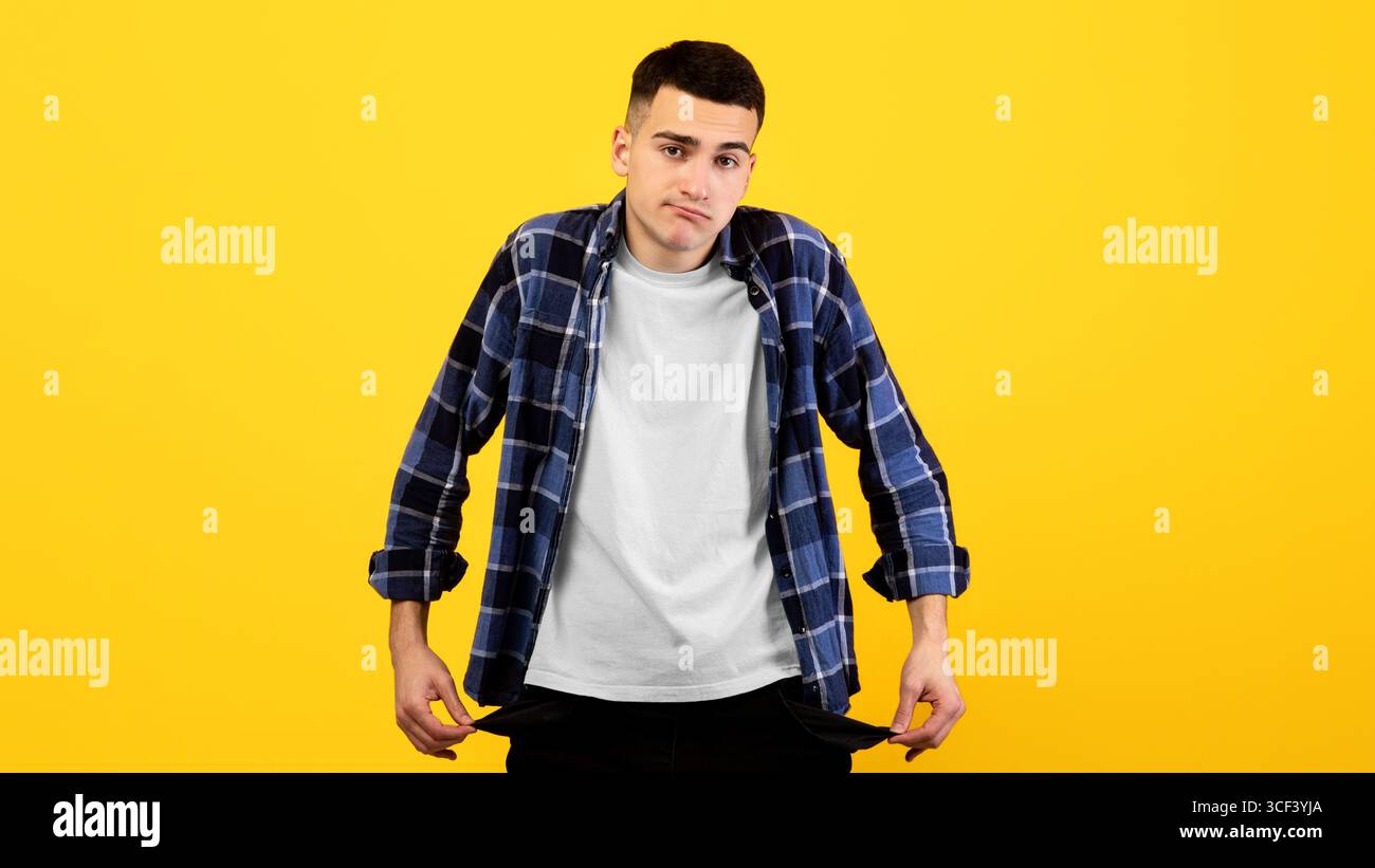 Un giovane ragazzo sconvolto che mostra tasche vuote sullo sfondo arancione dello studio Foto Stock