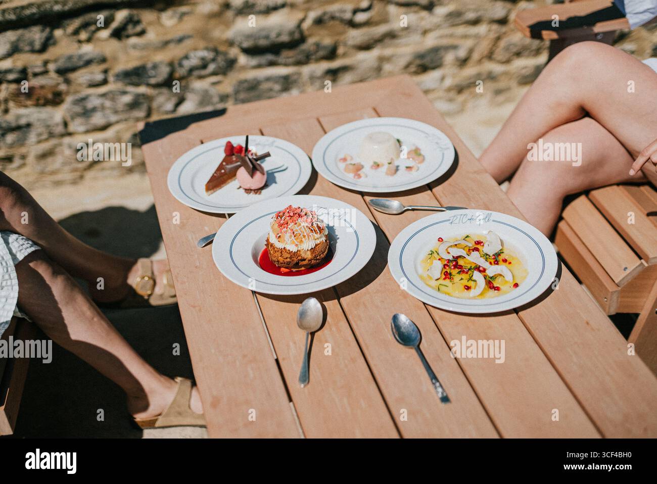 Un tavolo con quattro piatti di cibo, incluso un dessert con un condimento rosa. Due persone sono sedute al tavolo, una di loro indossa dei sandali Foto Stock