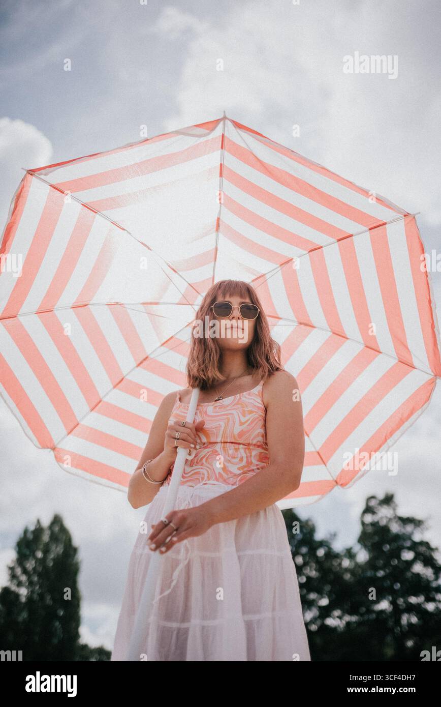 Una donna è in piedi sotto un grande ombrello a righe bianche e rosa. Indossa un vestito bianco e una canotta rosa. L'ombrello è aperto e fornisce Foto Stock Una donna è in piedi sotto un grande ombrello a righe bianche e rosa. Indossa un vestito bianco e una canotta rosa. L'ombrello è aperto e fornisce Foto Stock
