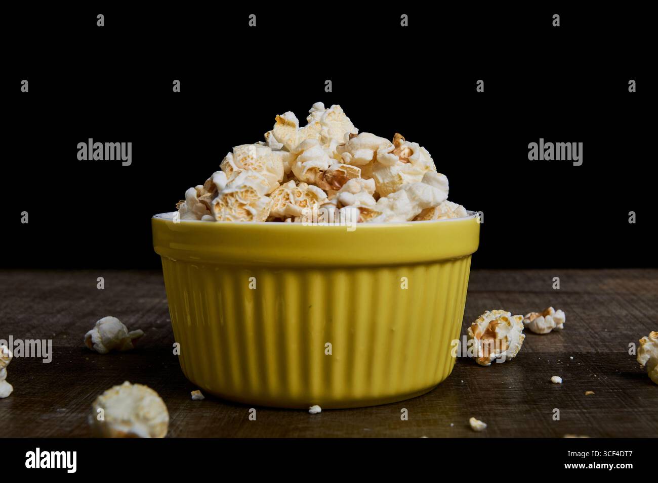 Recipiente giallo con popcorn su legno, fondo nero profondo. Composizione centrata, con minore dispersione. Ideale per la fotografia di prodotti o per alimenti minimalisti Foto Stock