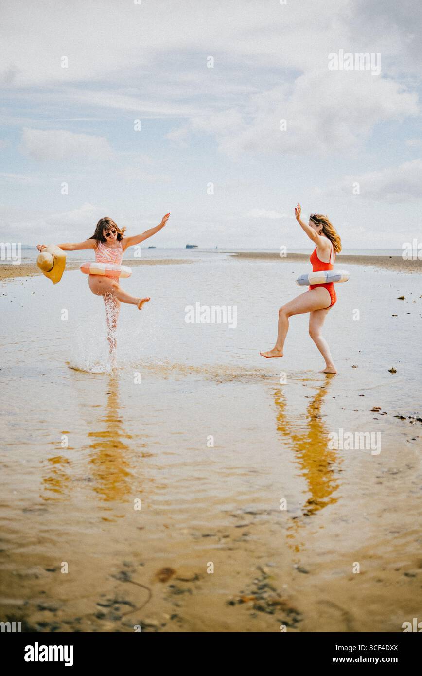 Due donne giocano in acqua, una delle quali indossa un costume da bagno rosso. L'acqua è calma e il cielo nuvoloso Foto Stock