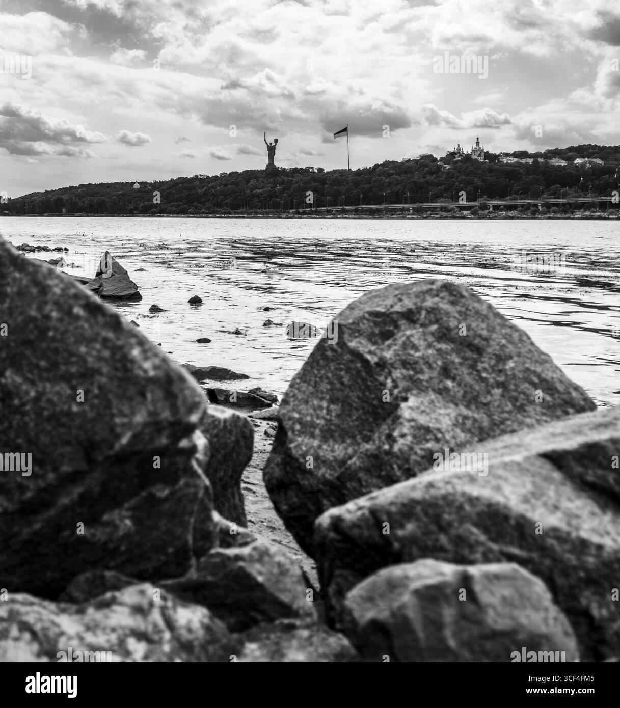 Immagine in bianco e nero di rocce sulla riva del fiume Dnipro con monumento madre Ucraina e bandiera Ucraina sullo sfondo sotto il cielo nuvoloso a Kiev Foto Stock