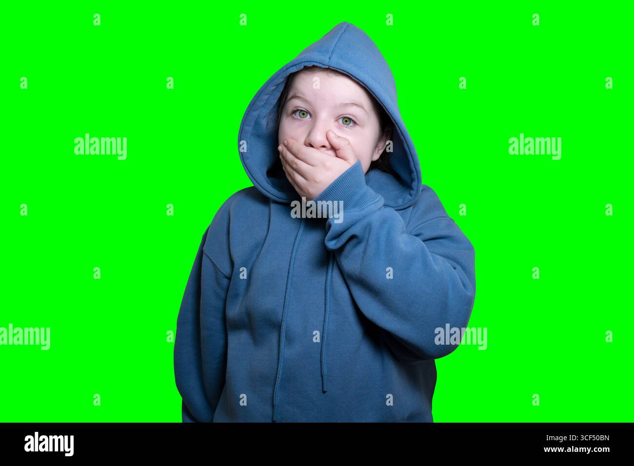 Una ragazza spaventata con gli occhi larghi in una felpa con cappuccio blu, che mette la mano sulla bocca, su uno sfondo verde per la chiave cromatica Foto Stock