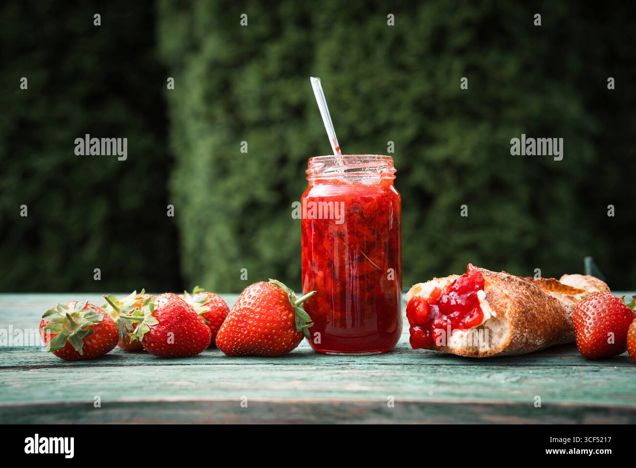 Marmellata di fragole fatta in casa in un barattolo con baguette fresca su un vecchio tavolo di legno all'aperto. Spalmabile a base di frutta fresca di stagione. Primo piano. Foto Stock