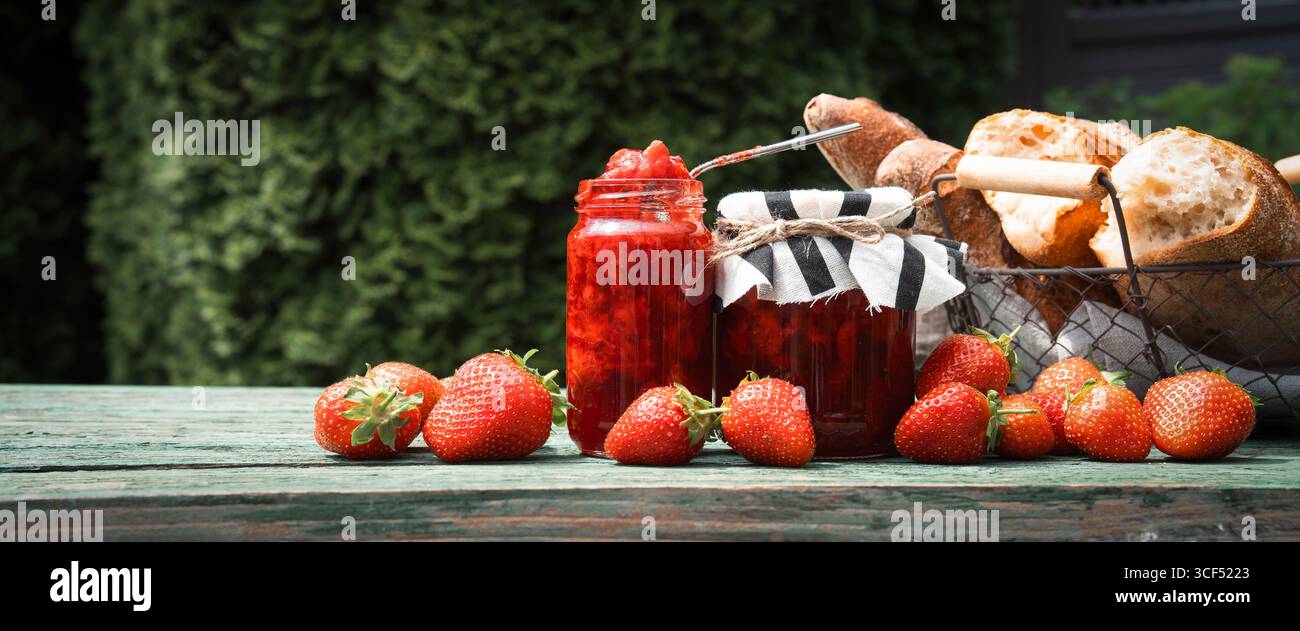 Marmellata di fragole fatta in casa in un barattolo con baguette fresca su un vecchio tavolo di legno all'aperto. Spalmabile a base di frutta fresca di stagione. Primo piano orizzontale. Foto Stock