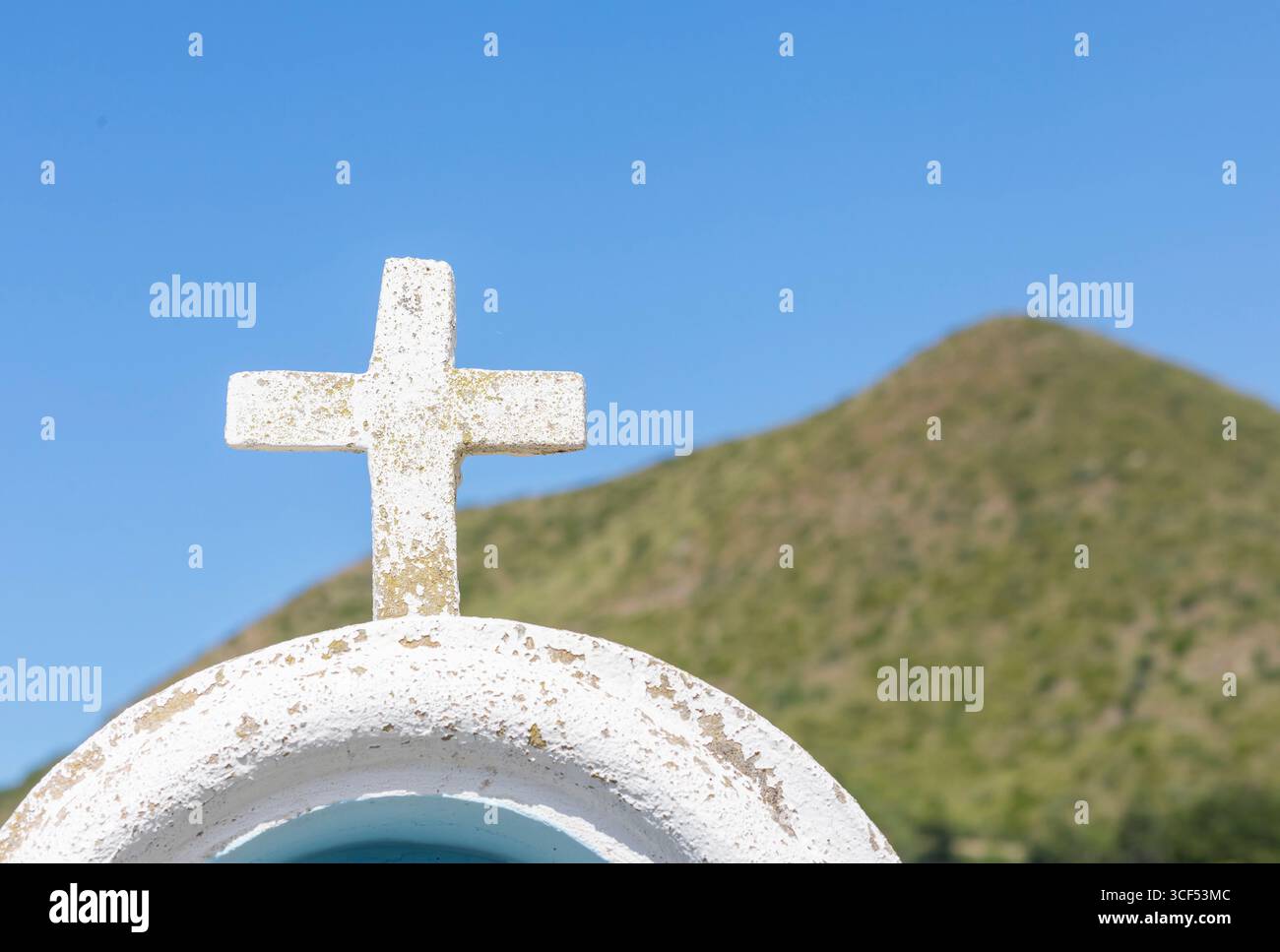 Croce bianca intemprata si erge in primo piano, affacciata su una lussureggiante collina verde sotto un vivace cielo blu, creando un paesaggio sereno e simbolico Foto Stock
