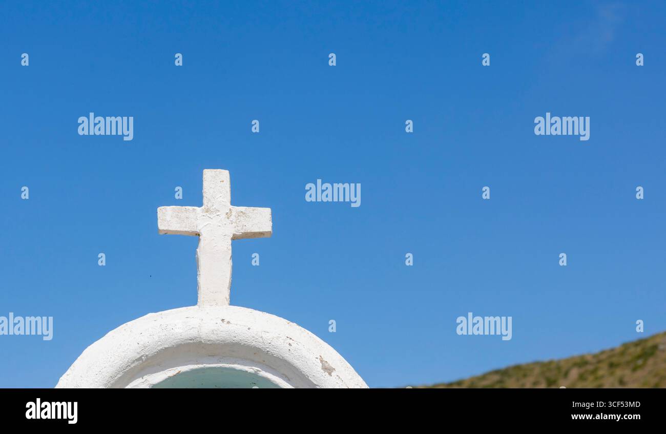 Croce bianca in piedi su un tetto imbiancato sotto un cielo azzurro, che rappresenta la religione, la fede, il cristianesimo e la spiritualità, con una collina sfocata sullo sfondo Foto Stock