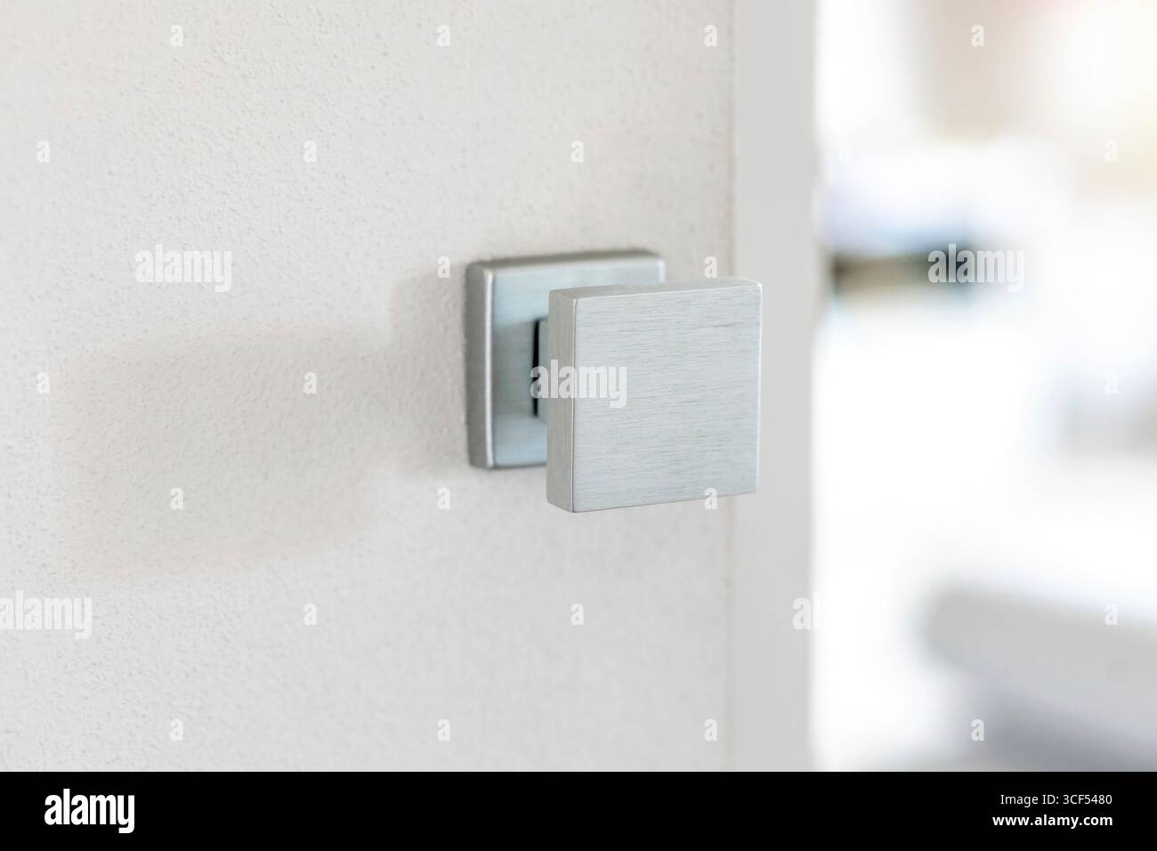 Primo piano di una moderna maniglia quadrata in metallo spazzolato montata su una porta bianca, leggermente socchiusa, che rivela uno spazio interno luminoso e sfocato, che enfatizza il design minimalista e l'arredamento contemporaneo della casa Foto Stock