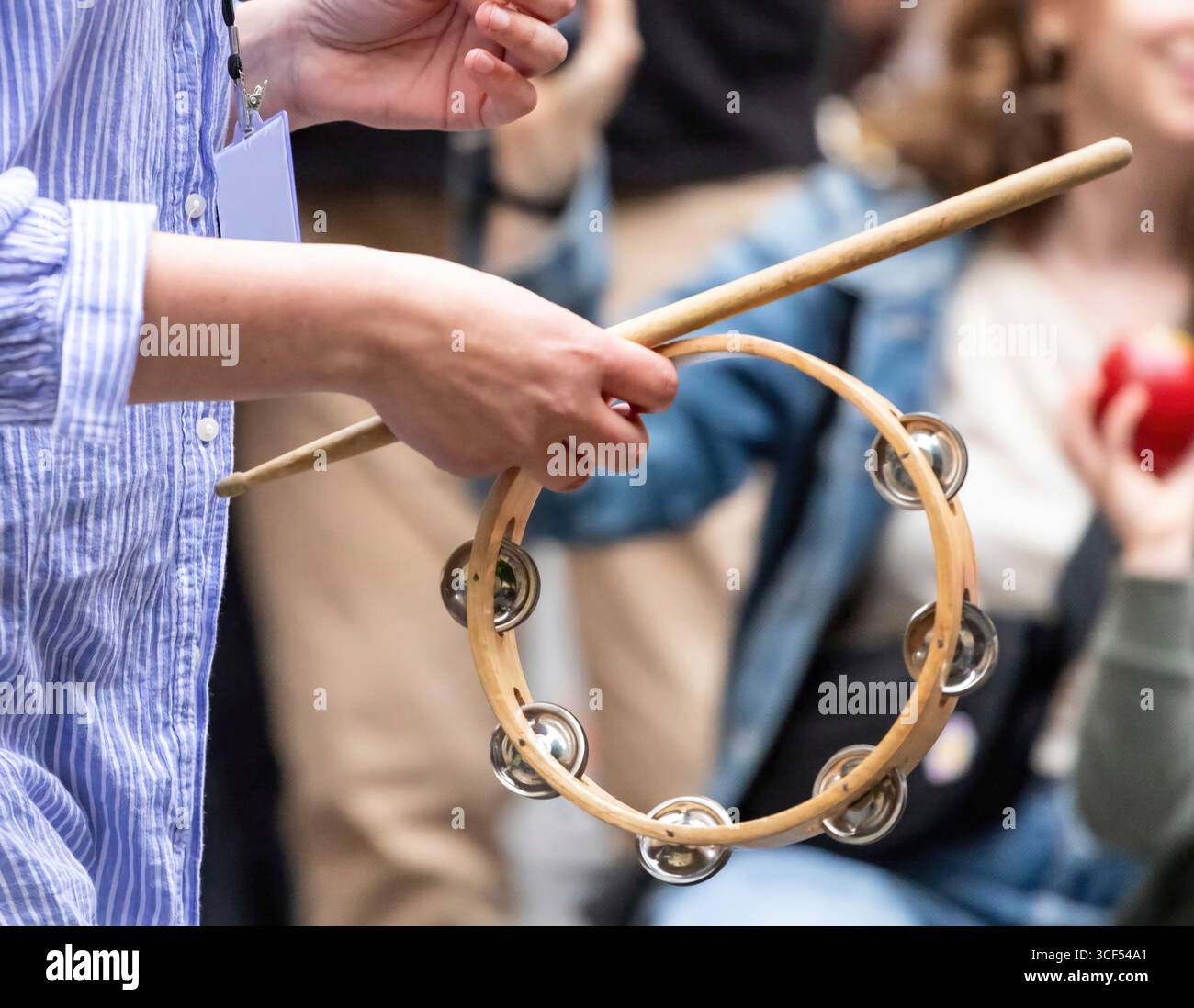 Primo piano delle mani di un musicista che suona un tamburello durante un concerto di strada, tenendo una bacchetta di legno e la cornice circolare con campane di metallo, creando un'atmosfera ritmica e festosa Foto Stock