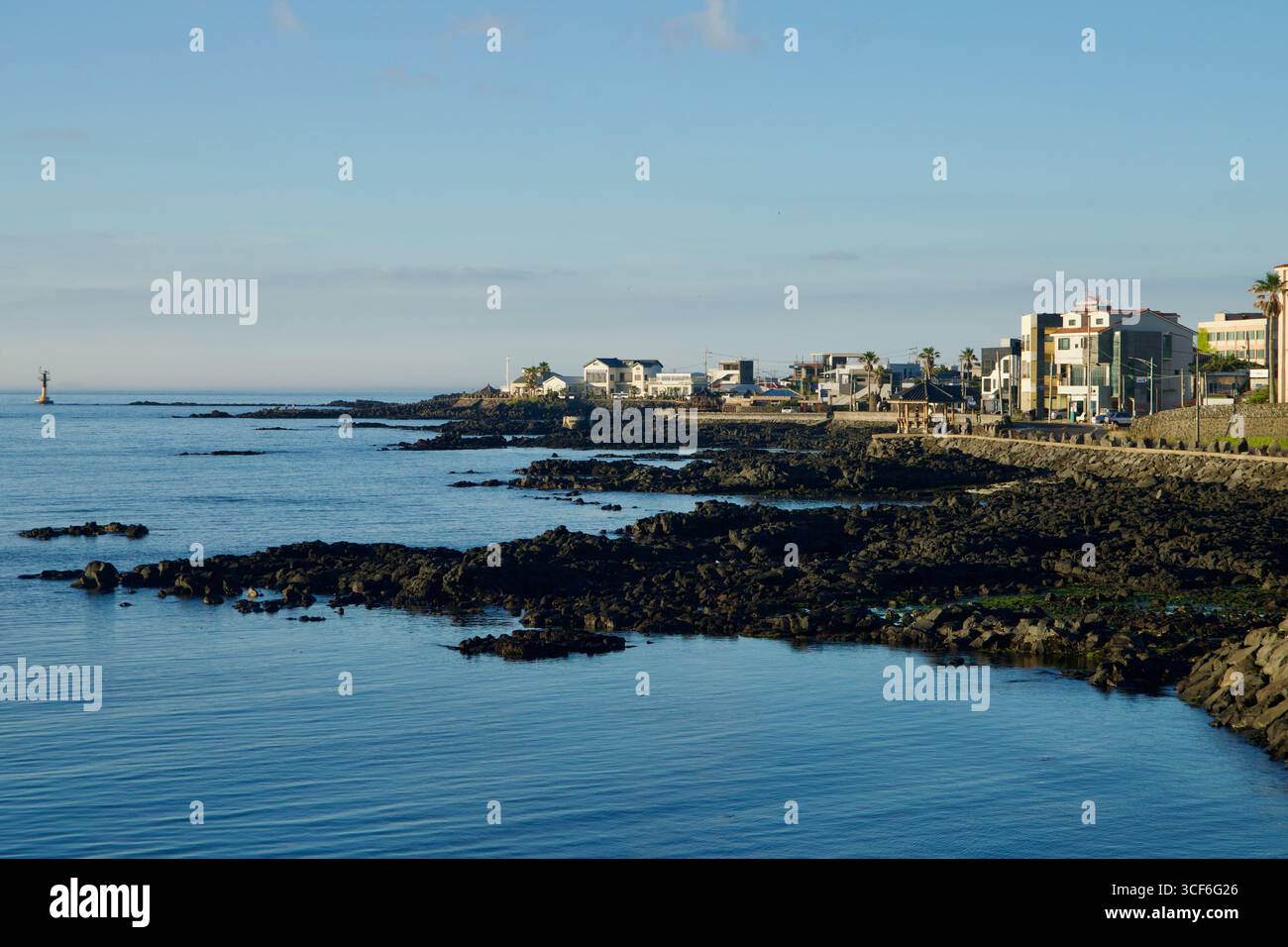 Affioramenti di lava basaltica circondano la tranquilla costa sotto il villaggio di Gwideok, con un faro lontano e moderne case sul mare che costeggiano la strada costiera di Jeju Foto Stock