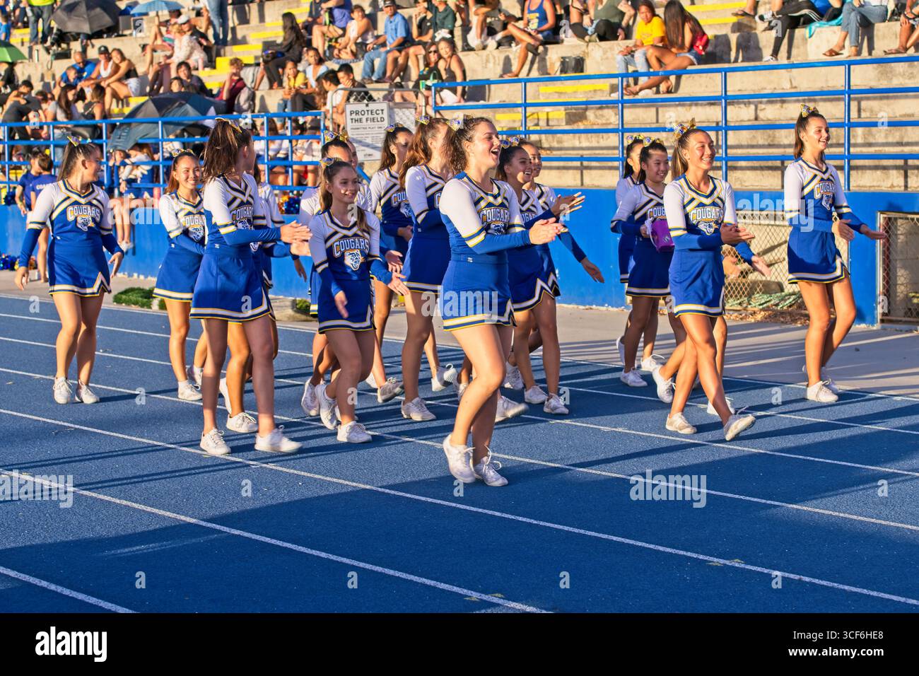 Le cheerleader con uniformi blu, gialle e bianche vibranti colpiscono pose dinamiche sotto il cielo dorato del tramonto di Hawai'i, irradiando orgoglio, spirito e unità. Foto Stock