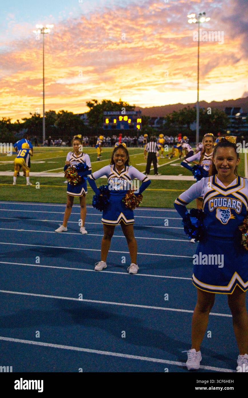 Le cheerleader con uniformi blu, gialle e bianche vibranti colpiscono pose dinamiche sotto il cielo dorato del tramonto di Hawai'i, irradiando orgoglio, spirito e unità. Foto Stock