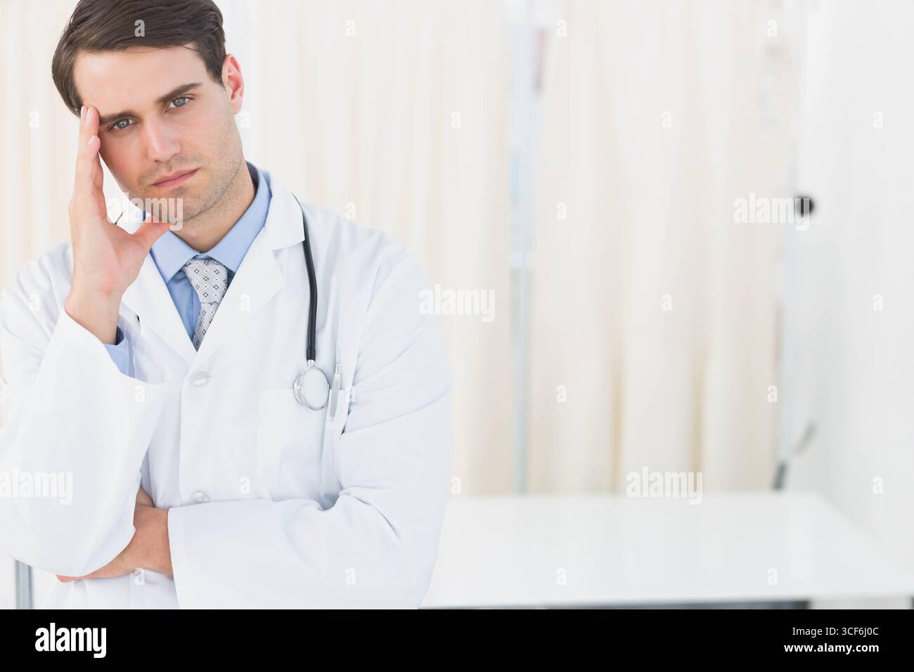 Medico di sesso maschile in piedi pensivamente in sala esame con camice da laboratorio accanto allo stetoscopio, spazio di copia Foto Stock