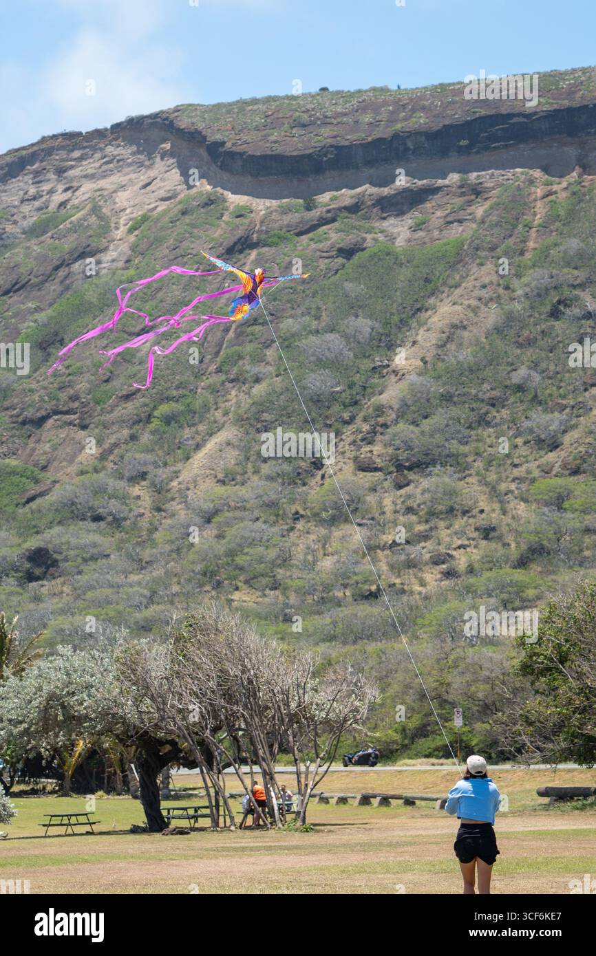 Una donna balla con il vento mentre fa volare un aquilone da sola su una spiaggia tropicale, lussureggianti montagne verdi che si innalzano dietro di lei in tranquilla serenità. Foto Stock
