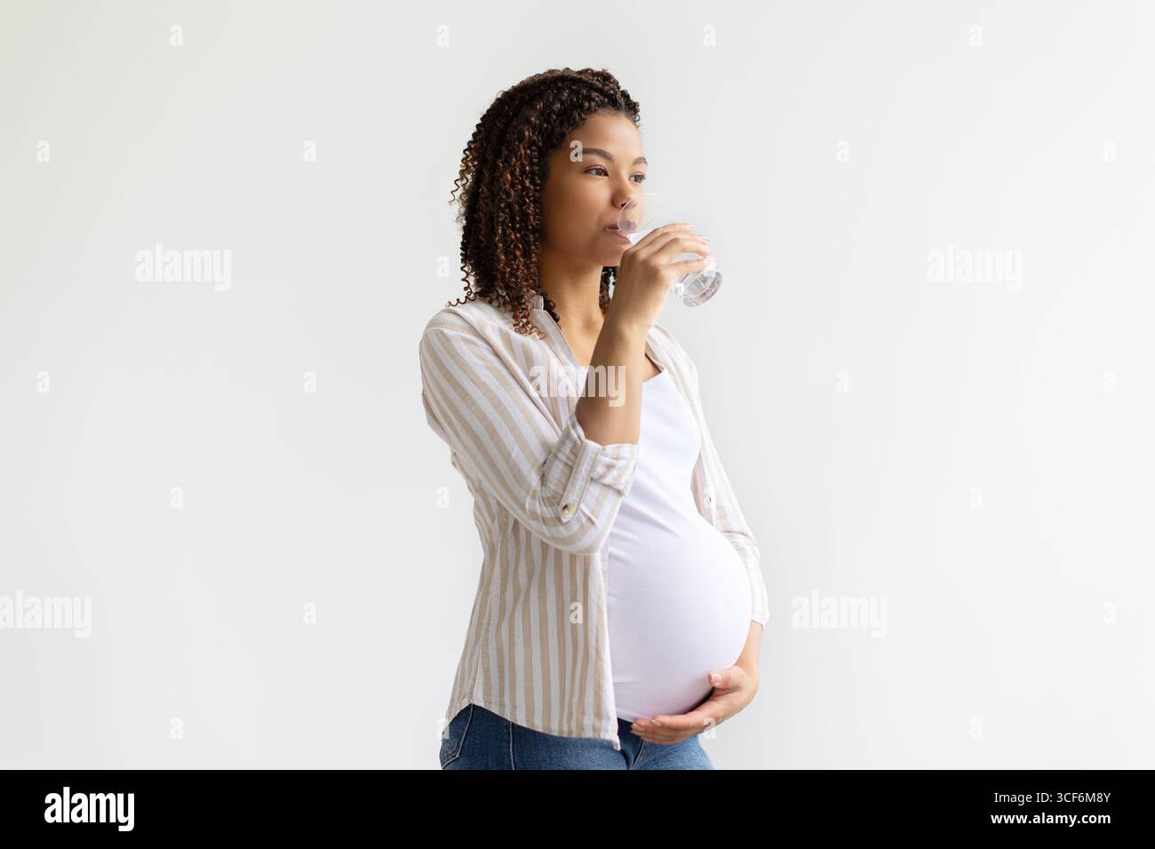 Donna afroamericana incinta che beve un bicchiere d'acqua e tocca la pancia Foto Stock