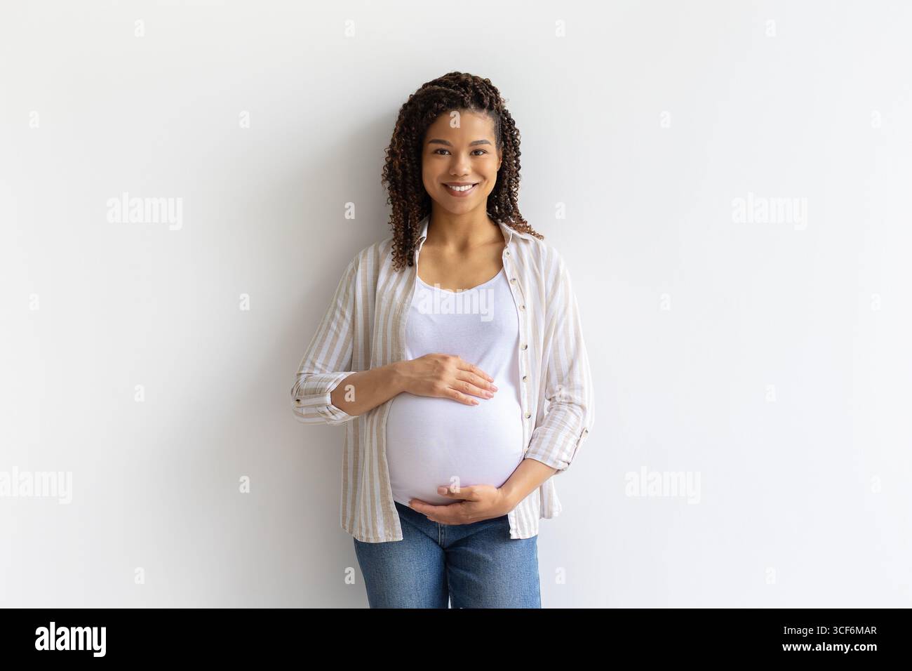 Sorridente donna nera incinta in piedi su sfondo bianco, tenendo la pancia con le mani Foto Stock