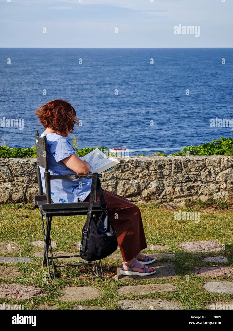 Una donna ama leggere un libro sul Paseo de San Pedro, circondato da erba e pietra, con una tranquilla barca che galleggia nel mare lontano. Foto Stock
