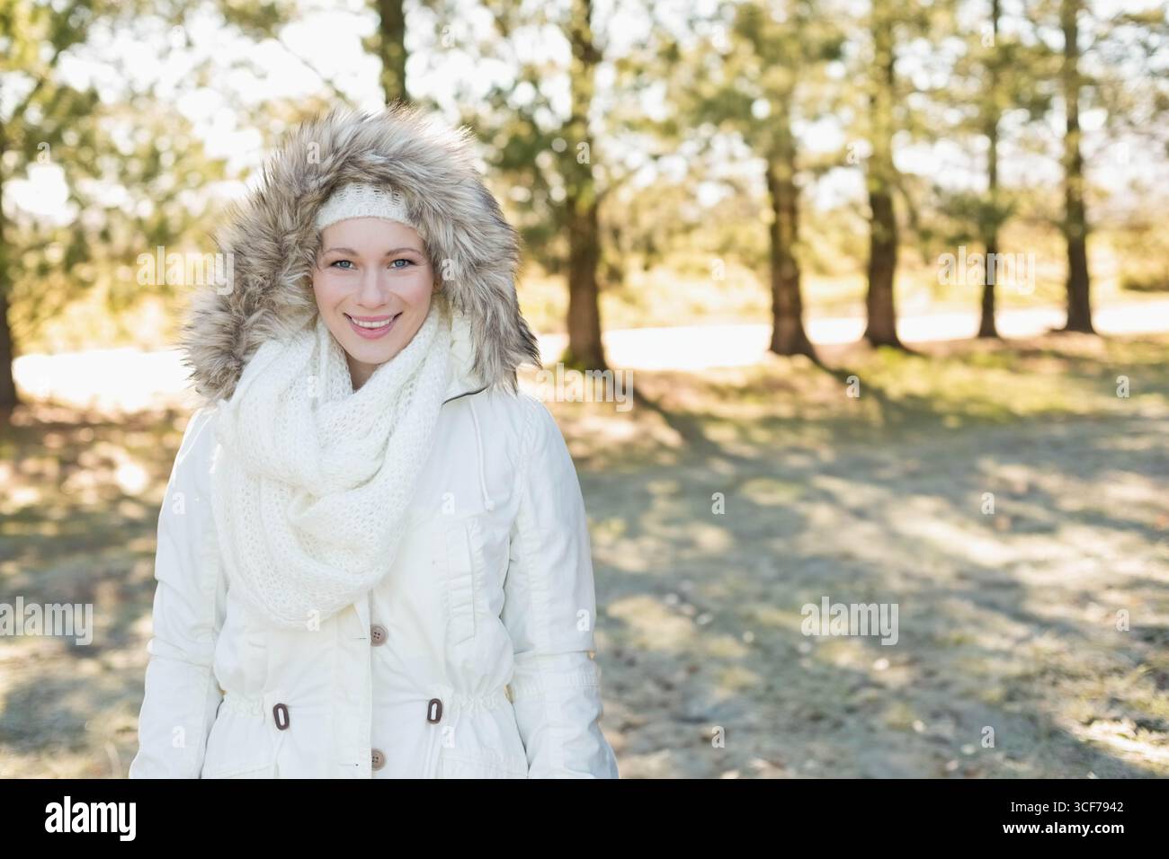 Donna in piedi in una pineta illuminata dal sole che indossa un parka bianco con finiture in pelliccia sintetica, sciarpa e fascia Foto Stock