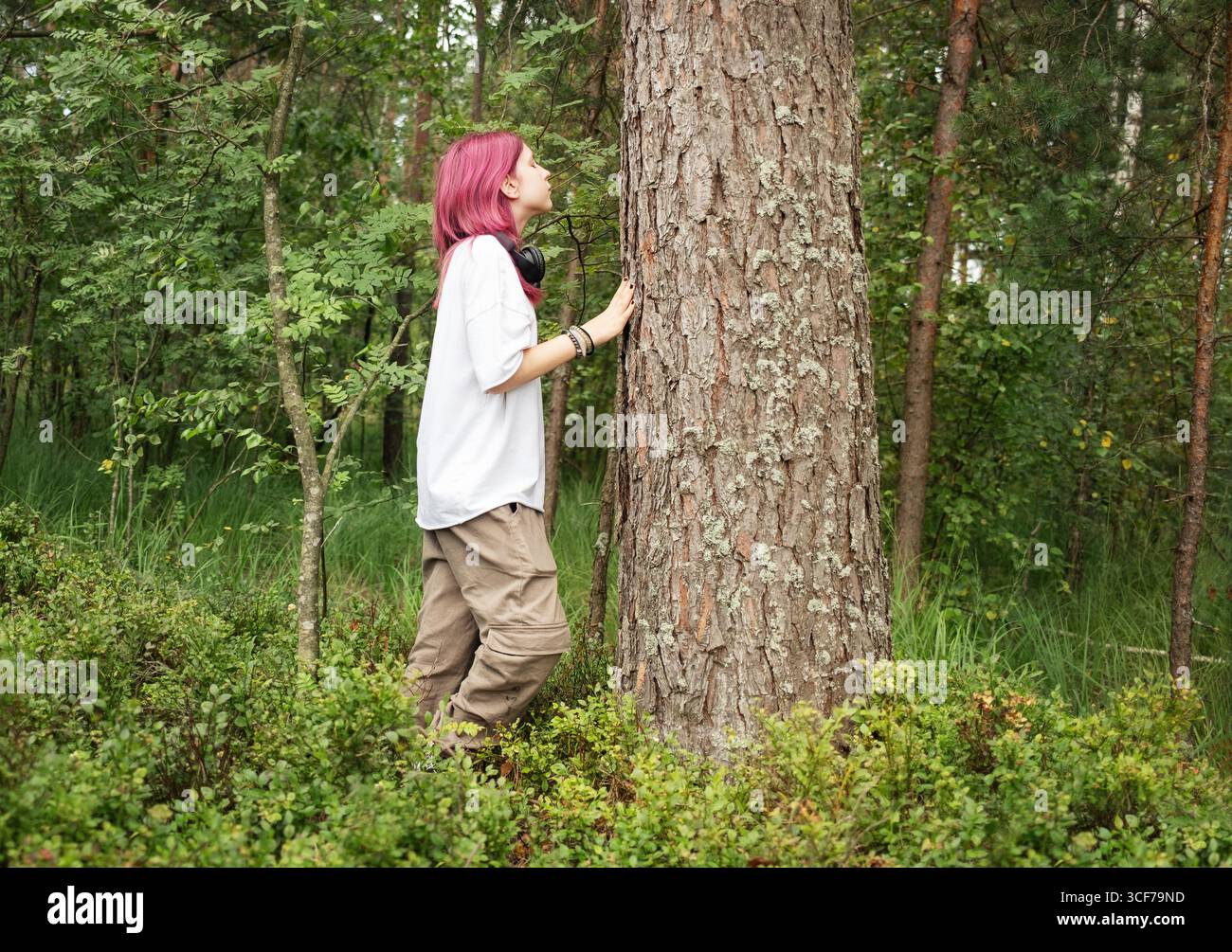 L'escursionista con i capelli rosa abbraccia un grande tronco di albero in una lussureggiante foresta verde, che collega con la natura Foto Stock