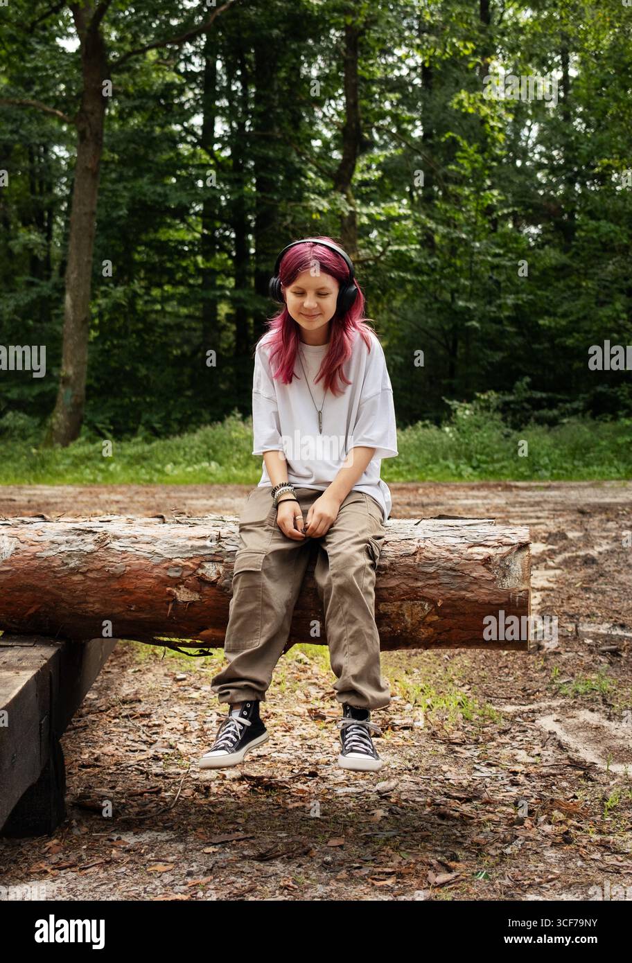 Adolescente con i capelli rosa che ascolta la musica in natura, seduto su un tronco d'albero Foto Stock