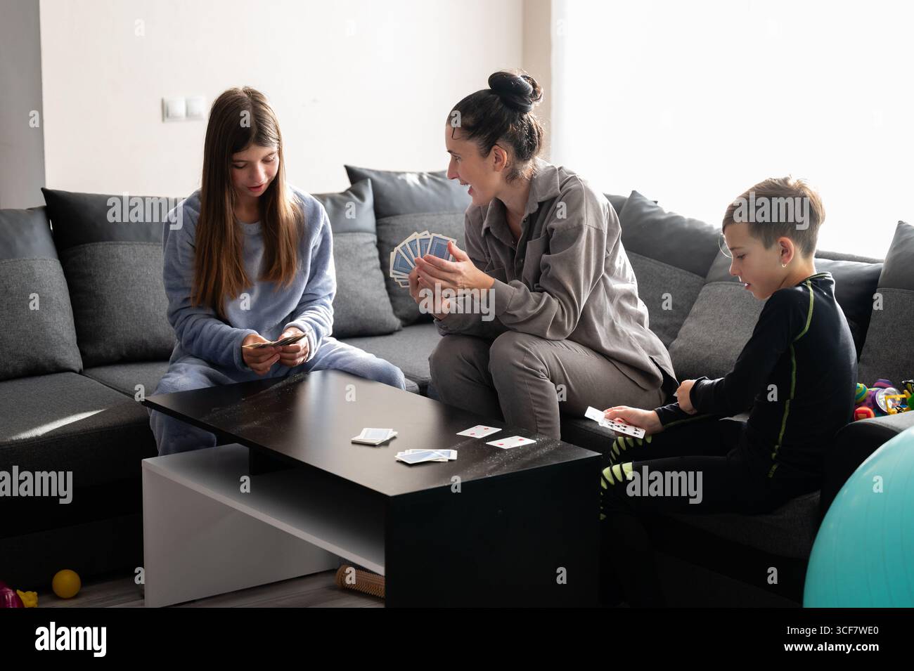 Due bambini e un adulto partecipano a un vivace gioco di carte su un divano moderno, pieno di gioia ed eccitazione in un luminoso soggiorno pieno di Foto Stock