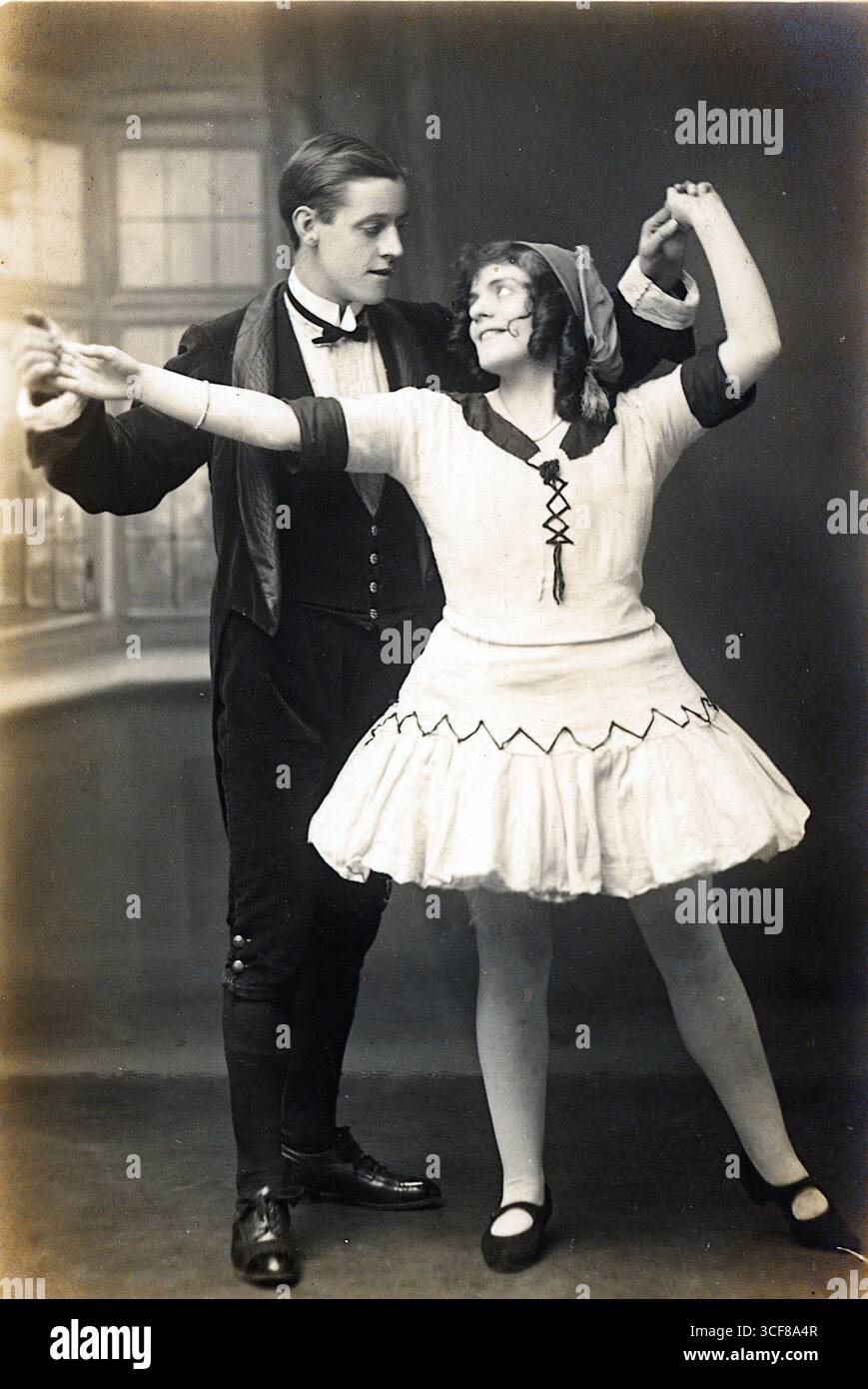 Questa cartolina dei primi anni del XX secolo, pubblicata da T. A. Stephenson di South Queen Street, Morley, cattura Deane e Hird nel bel mezzo della danza in un momento drammatico. L'uomo indossa un abito scuro con cravatta e scarpe con bottoni alti, mentre la donna indossa un costume da balletto con gonna svasata, collant e velo. La loro posa aggraziata, con le mani unite e le braccia allungate, evoca l'eleganza edoardiana del palcoscenico. Ambientata all'interno davanti a una finestra a più riquadri, l'immagine unisce luce naturale e formalità da studio, offrendo uno sguardo alla moda del periodo, alle performance e alla cultura da cartolina. Foto Stock