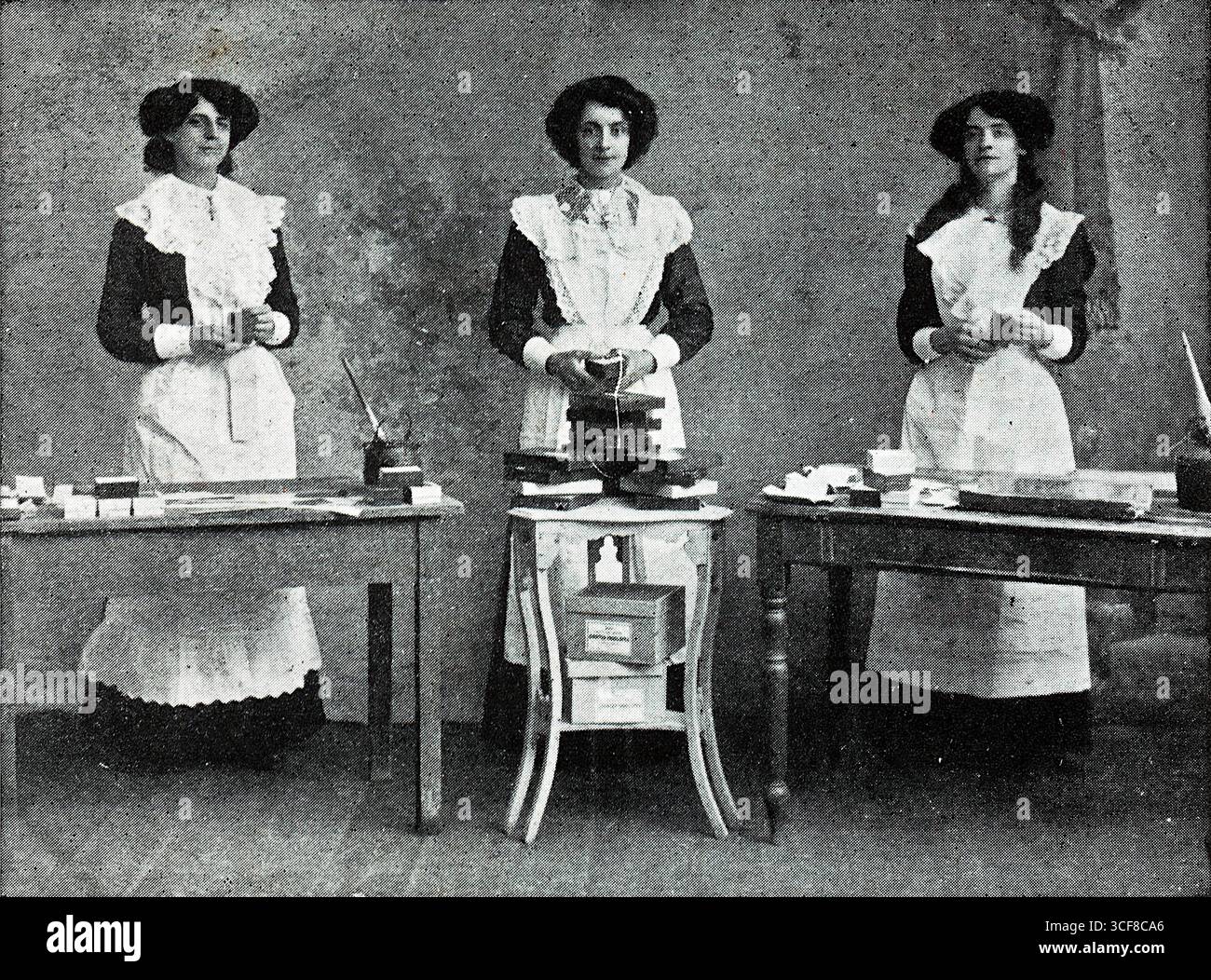 Dolly, Maud e Agnes, noti come i famosi boxmaker cantanti, si trovano dietro due tavoli da lavoro, vestiti con lunghi abiti scuri abbinati, grembiuli bianchi e cappelli evocativi dell'abbigliamento di fabbrica dei primi anni del XX secolo. Le loro espressioni articolate e le caselle ordinate suggeriscono sia l'abilità manuale che la presenza performativa. Probabilmente allestita in un'officina, l'immagine cattura l'intersezione tra lavoro e intrattenimento. Intorno al 1910, questa fotografia riflette il fascino edoardiano per gli atti di novità e l'identità teatrale della classe operaia, mescolando l'industria con il canto e la cameratismo. Foto Stock