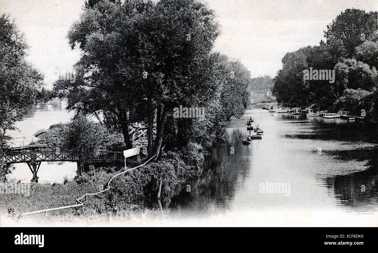 Questo paesaggio fluviale monocromatico cattura il fascino tranquillo del braccio e dell'isola di Migneaux di Poissy, probabilmente all'inizio del XX secolo. Una passerella in legno sbircia attraverso fitte foglie sulla sinistra, mentre un sentiero stretto o una tubatura curva lungo la riva ombreggiata. Le piccole barche riposano lungo la riva destra sotto un baldacchino frondoso, suggerendo svago e quiete. La composizione bilancia il recinto naturale con una sottile presenza umana. Pubblicata da Léon Levy, l'immagine evoca una pausa contemplativa in un paesaggio dolcemente abitato. Foto Stock