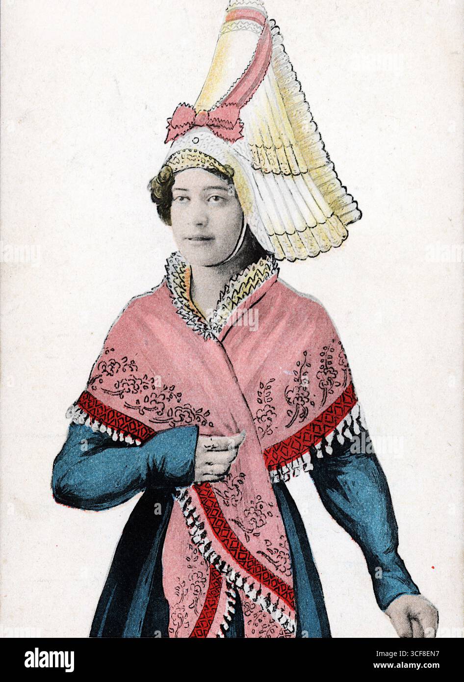 Questo suggestivo copricapo evoca l'eleganza cerimoniale della Normandia durante il regno di Enrico II (1547-1559). L'alto e strutturato cofano in pizzo, a strati di bianco, rosa e giallo, suggerisce una variazione regionale all'interno della moda rinascimentale francese, probabilmente adattato da interpretazioni folcloristiche successive. Abbinato a un abito blu a maniche lunghe e a uno scialle rosa con frange ornato da motivi floreali rossi e neri, l'insieme fonde riverenza storica con tradizione stilizzata. L'immagine cattura l'eredità duratura dell'abito normanno, dove l'orgoglio civico e gli echi cortesi convergono. Foto Stock