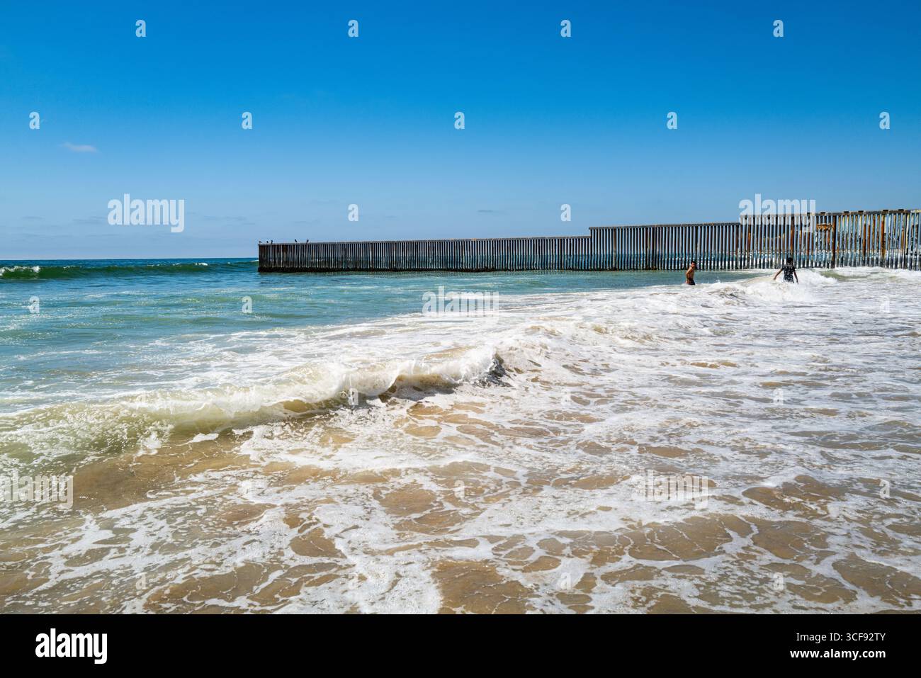 Vista lontana di giovani uomini nell'Oceano Pacifico vicino alla fine del muro di confine tra California e Messico che termina nell'oceano al Playas de Tijuana Foto Stock