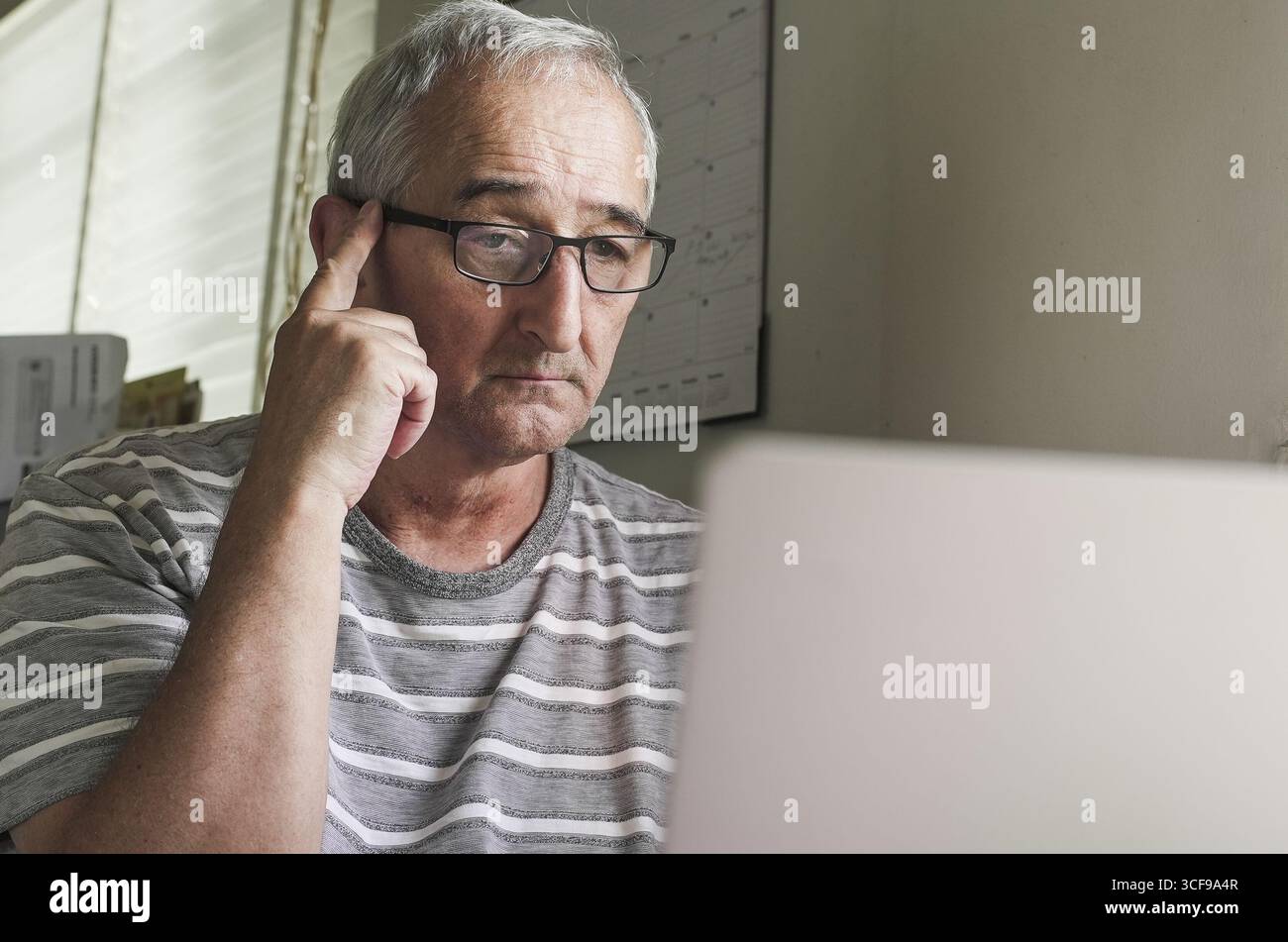 Un uomo anziano siede a una scrivania e si concentra su un notebook in una stanza ben illuminata. Ha i capelli grigi e indossa gli occhiali, apparendo contemplativo mentre tocca il suo per Foto Stock